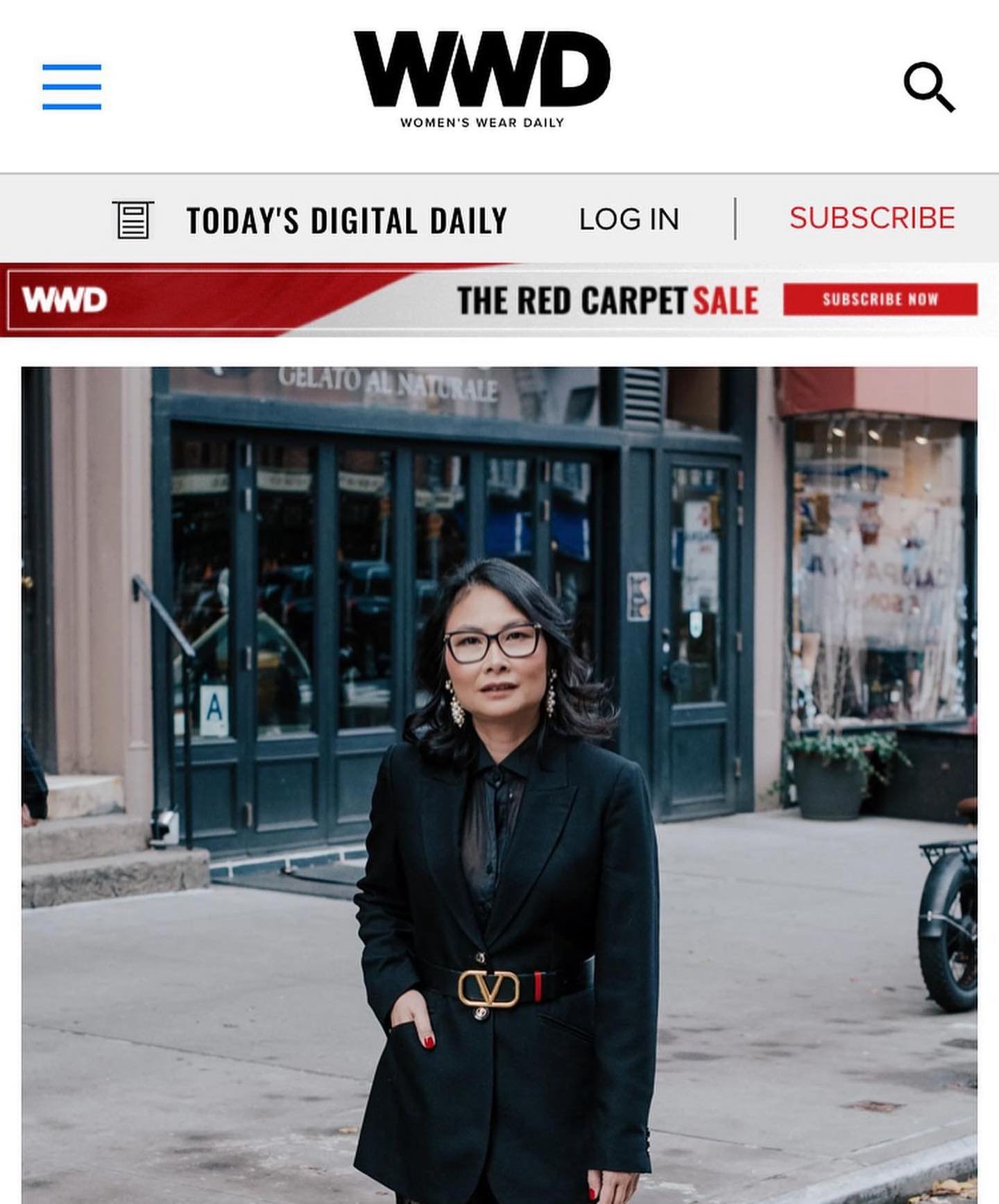 A woman in a black suit with a Valentino belt and glasses, standing on a city sidewalk in front of a storefront with a sign that says 'Gelato Al Naturale'. She has dark hair and earrings, and a motorcycle is partially visible to the right.
