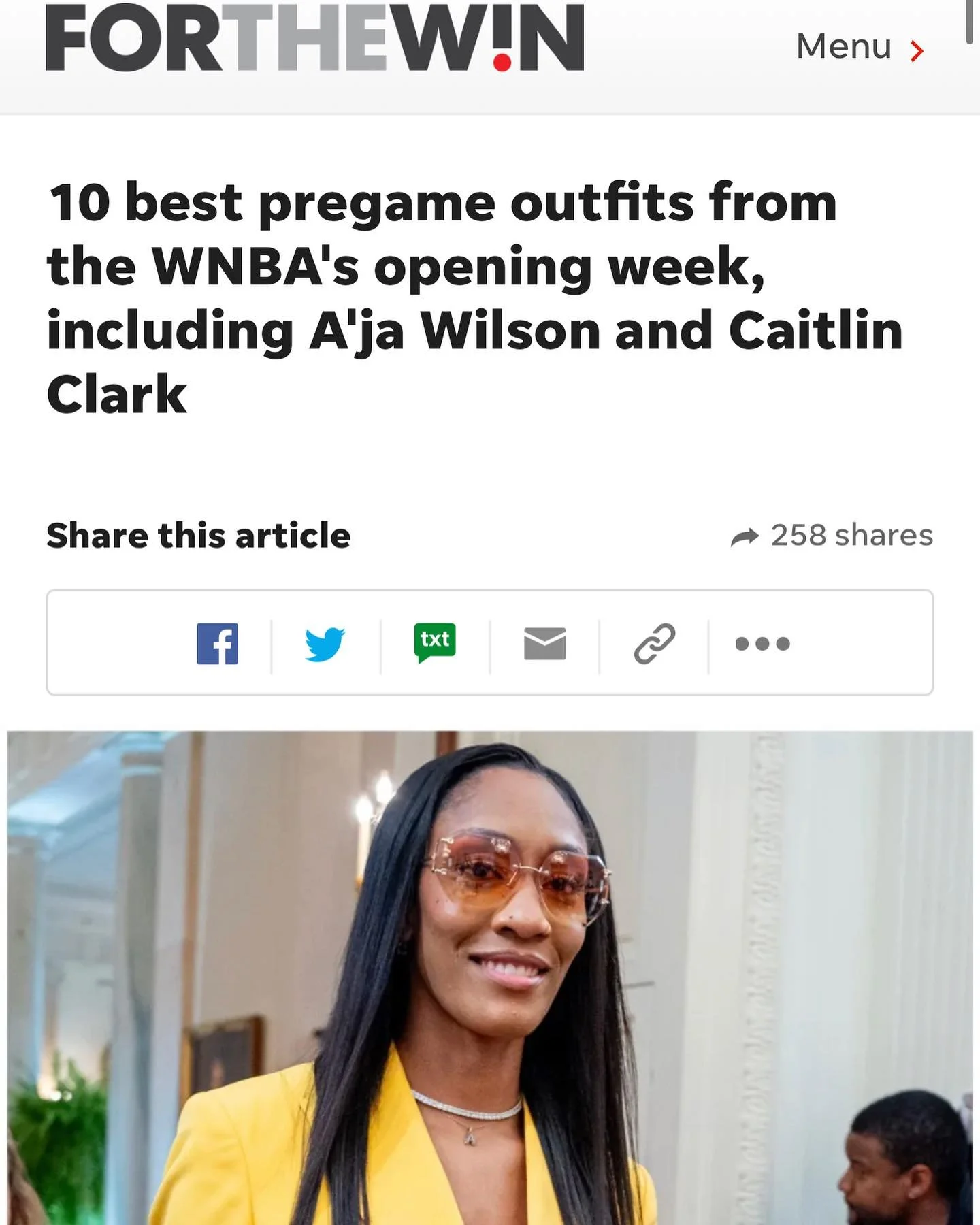 A woman with long black hair wearing sunglasses, a yellow blazer, and a necklace, smiling indoors.