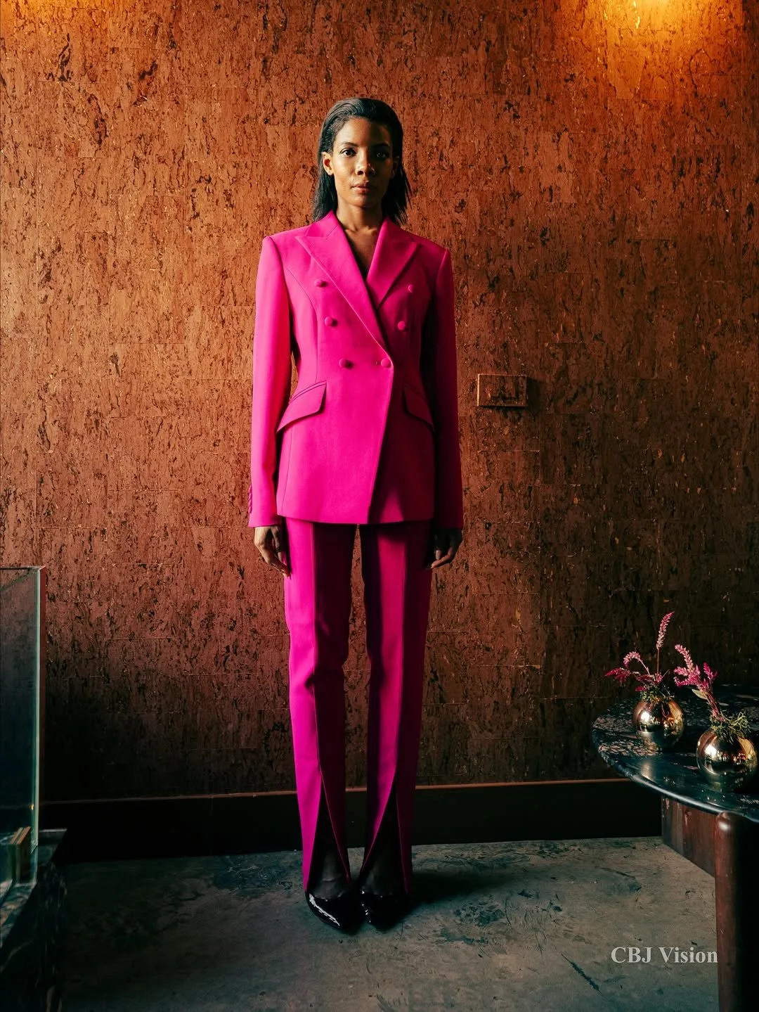 A woman wearing a bright pink pantsuit with black shoes, standing against a brown textured wall with a floral arrangement in metallic vases on a black table.