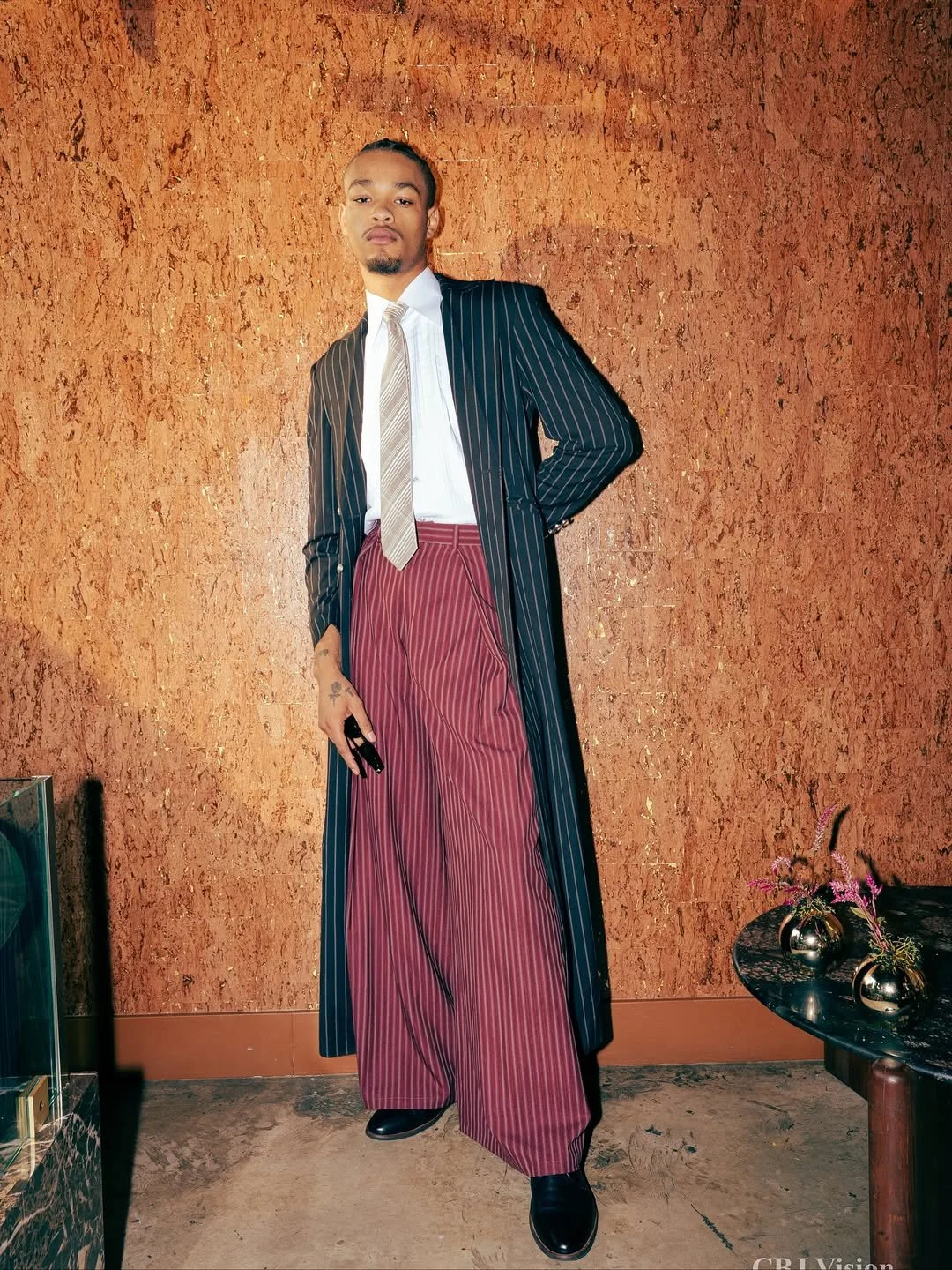 A young man wearing a black striped suit jacket, white shirt, light beige tie, and red wide-leg pinstripe pants, standing against a textured brown wall, with a small black table holding silver vases with flowers nearby.
