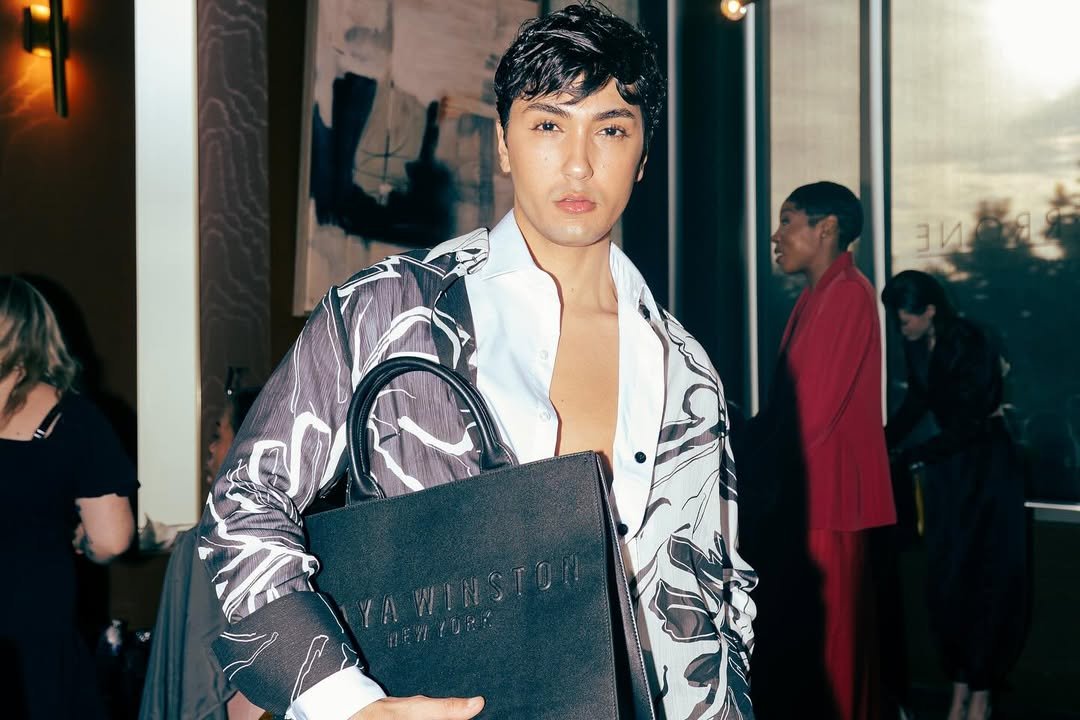 A young man with dark hair and wearing a patterned jacket over a partially open white shirt, holding a black Yawinston New York shopping bag, standing indoors in a retail or event space.