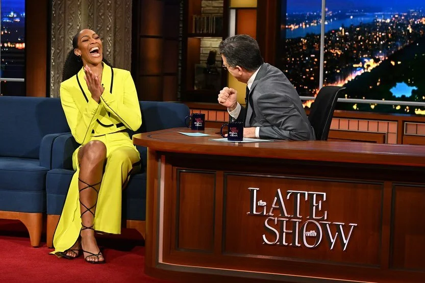 Tina Knowles laughing and clapping while sitting on a blue couch with a talk show host at a desk that says "The Late Show."