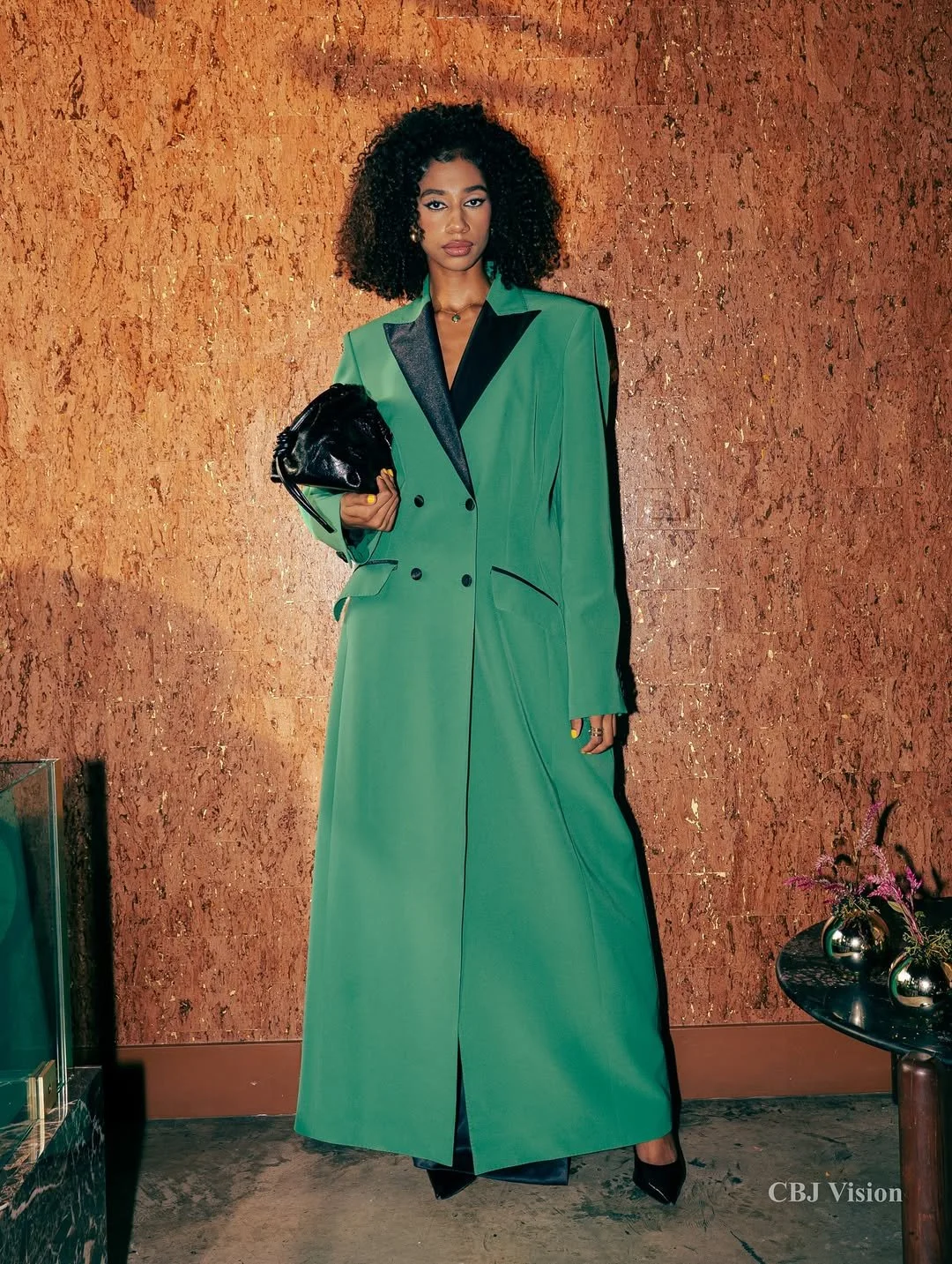 A woman with curly hair stands against a textured brown wall, wearing a long green blazer with black accents, holding a black handbag, with black shoes visible at the bottom.