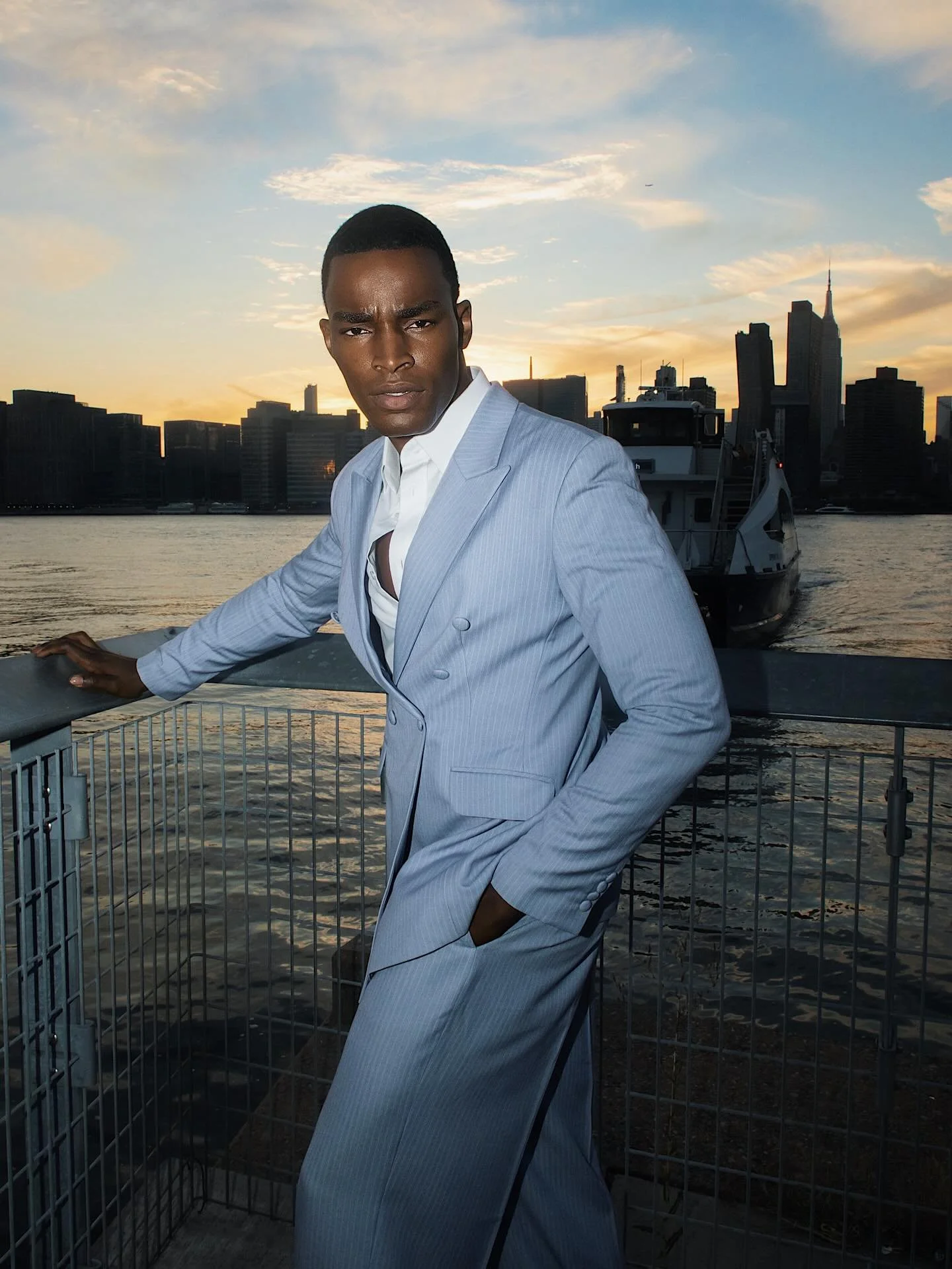 A man in a light gray pinstripe suit leaning on a railing by the water with a city skyline and sunset in the background.