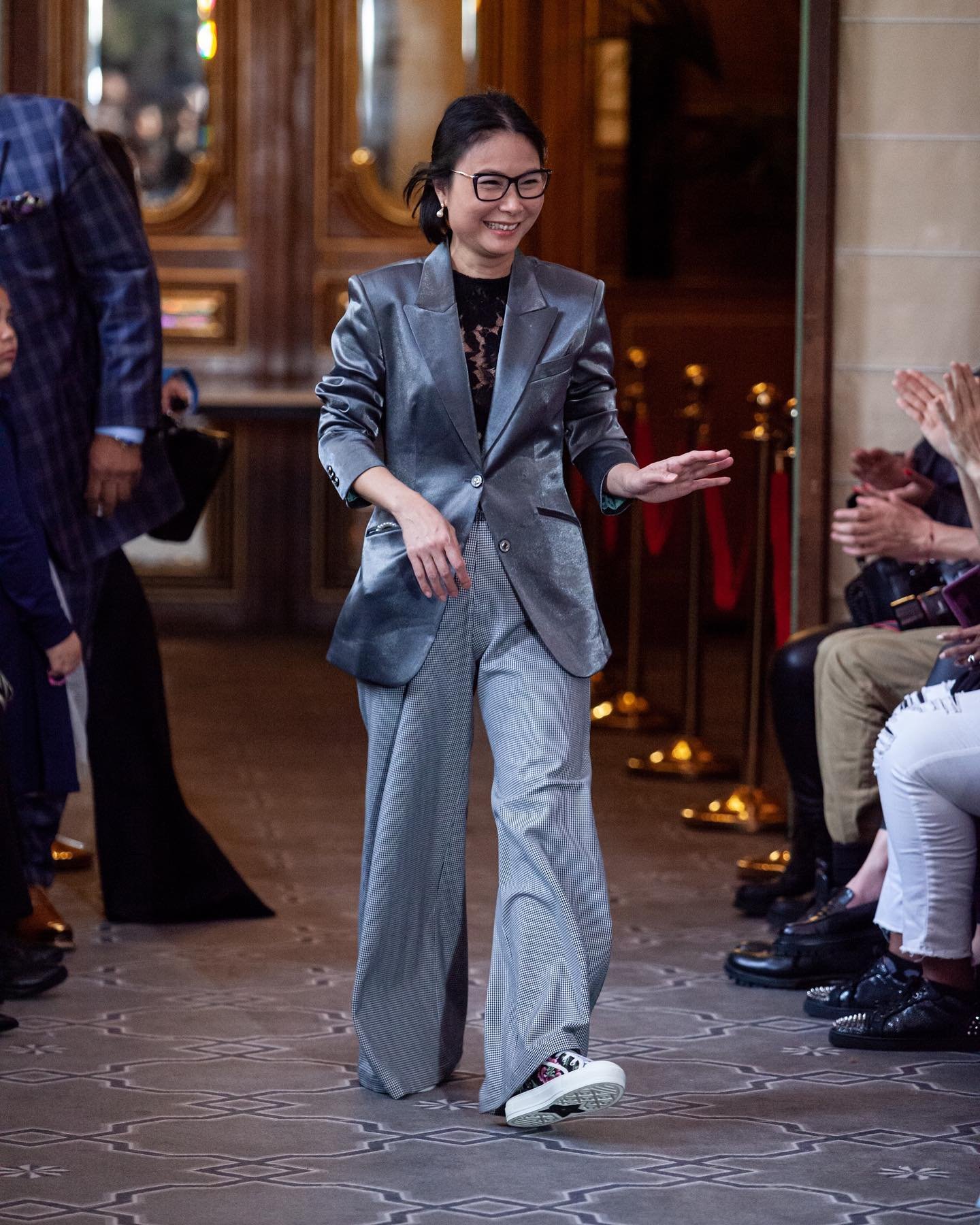 Woman in silver blazer and gray wide-leg trousers walking on a runway, smiling, in front of seated audience clapping.
