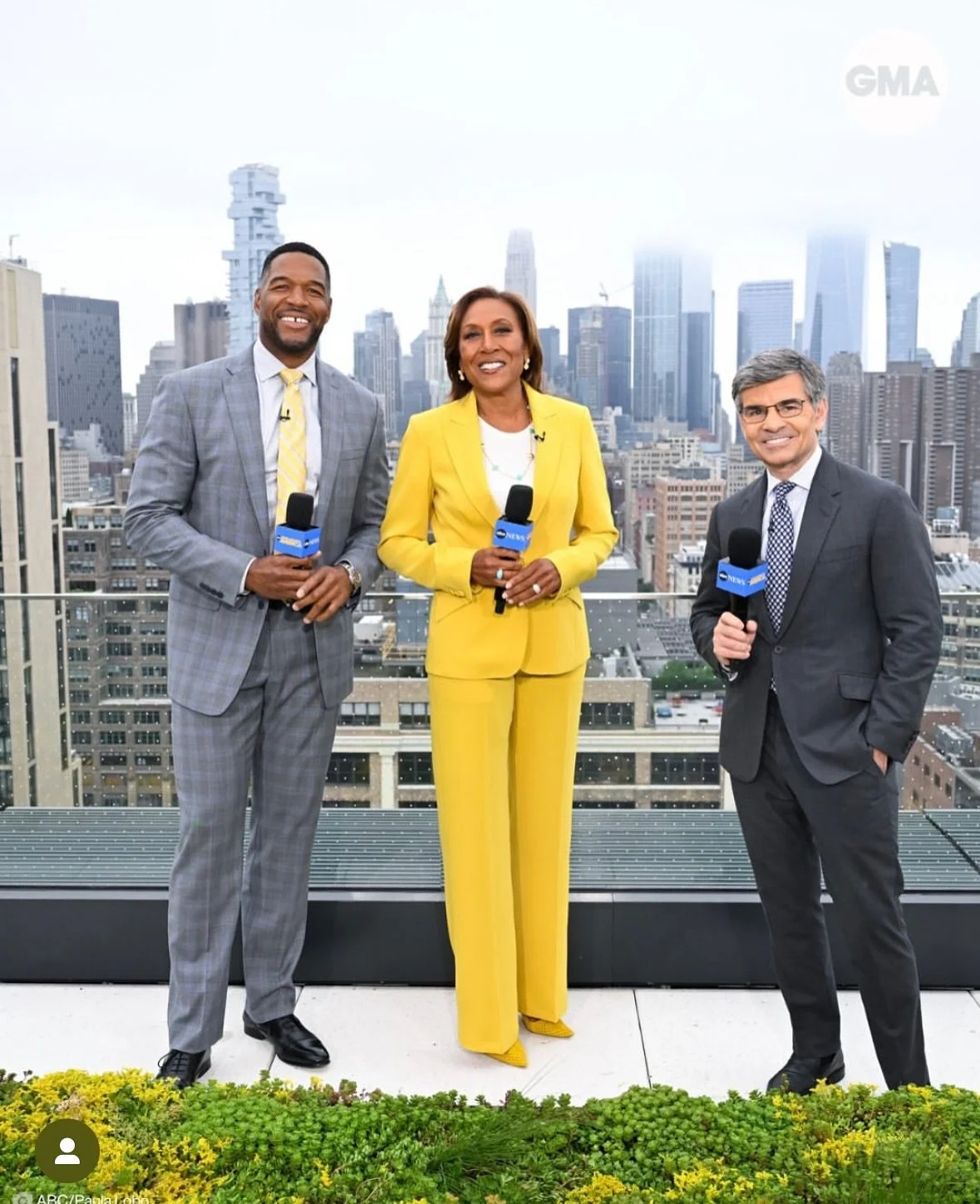 Three news anchors standing on a rooftop with New York City skyline in the background, holding microphones and smiling.