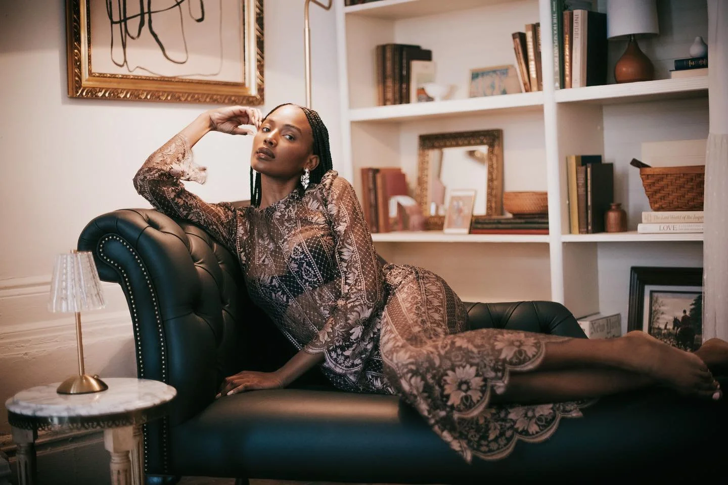 A woman with braided hair and dark lipstick lying on a black leather tufted chaise lounge in a well-decorated room with a bookshelf, framed artwork, a mirror, and a small round marble-top side table with a lamp.