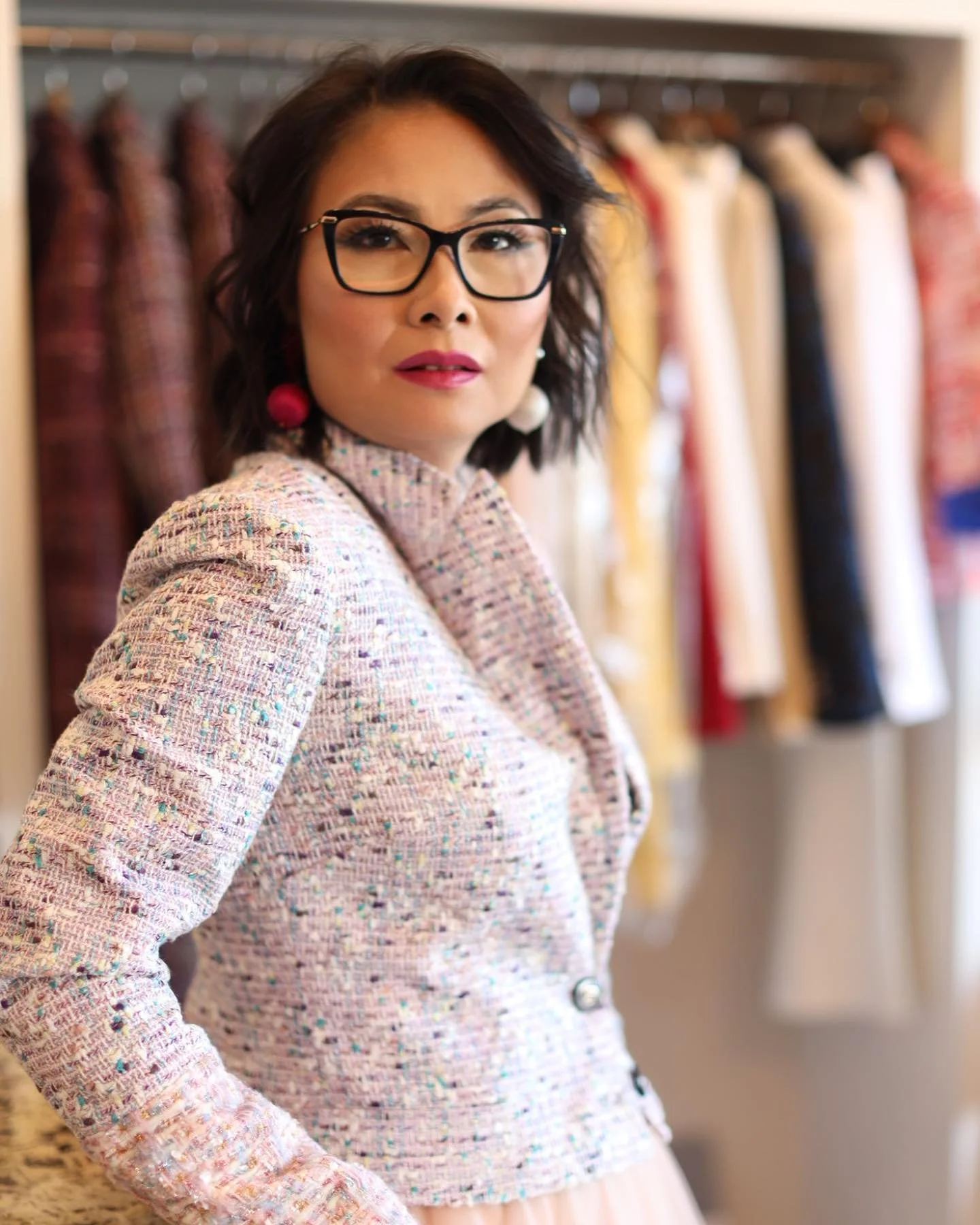 A woman with black hair, glasses, pink lipstick, and earrings stands in front of a clothing rack with colorful dresses.
