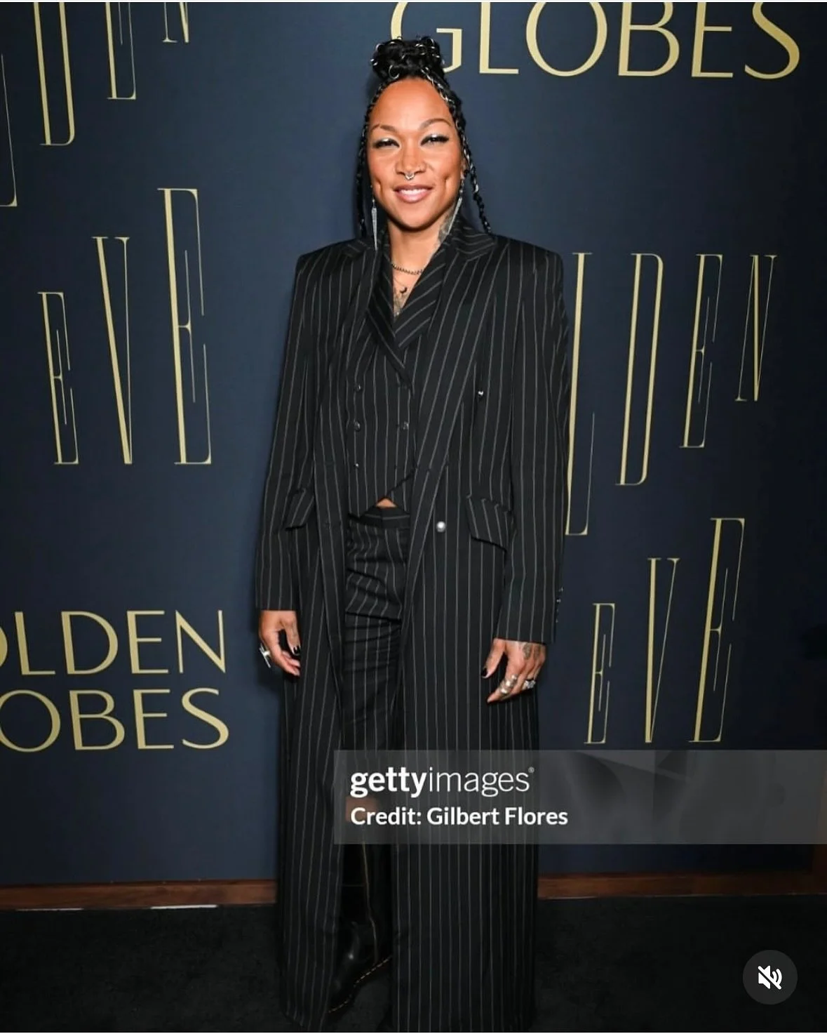 A woman wearing a black pinstripe suit standing in front of a dark backdrop with the words "GOLDEN GLOBES" written repeatedly in large gold letters.