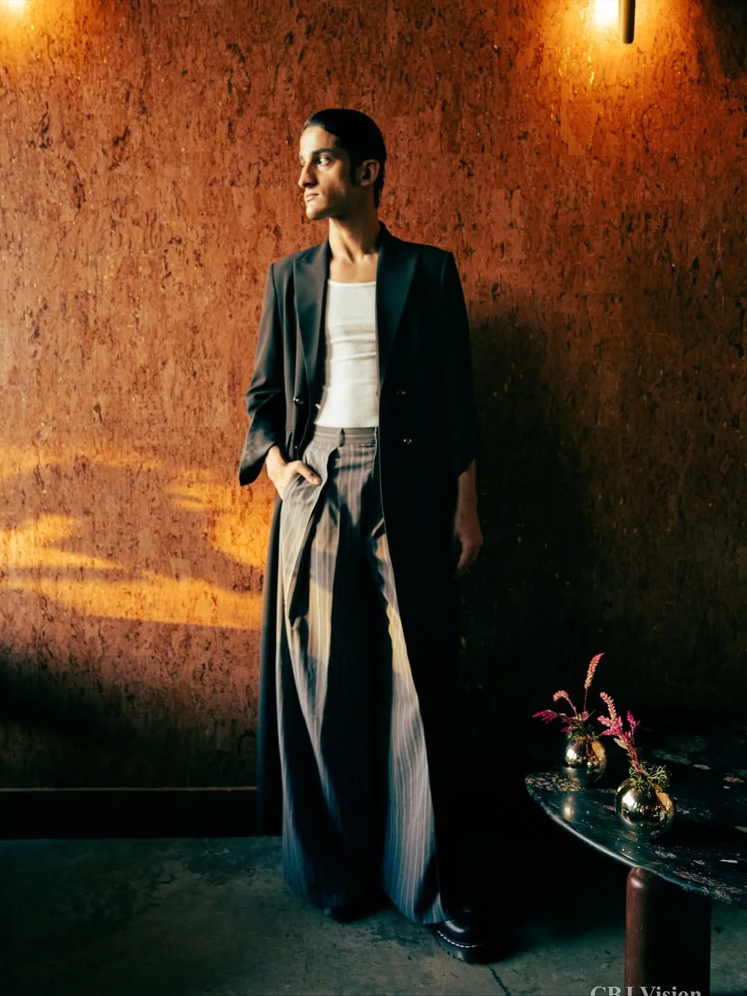 Person standing against a textured, warm-toned wall, wearing a black blazer, white top, and wide-legged striped pants, with their left hand in their pocket and looking to the left.