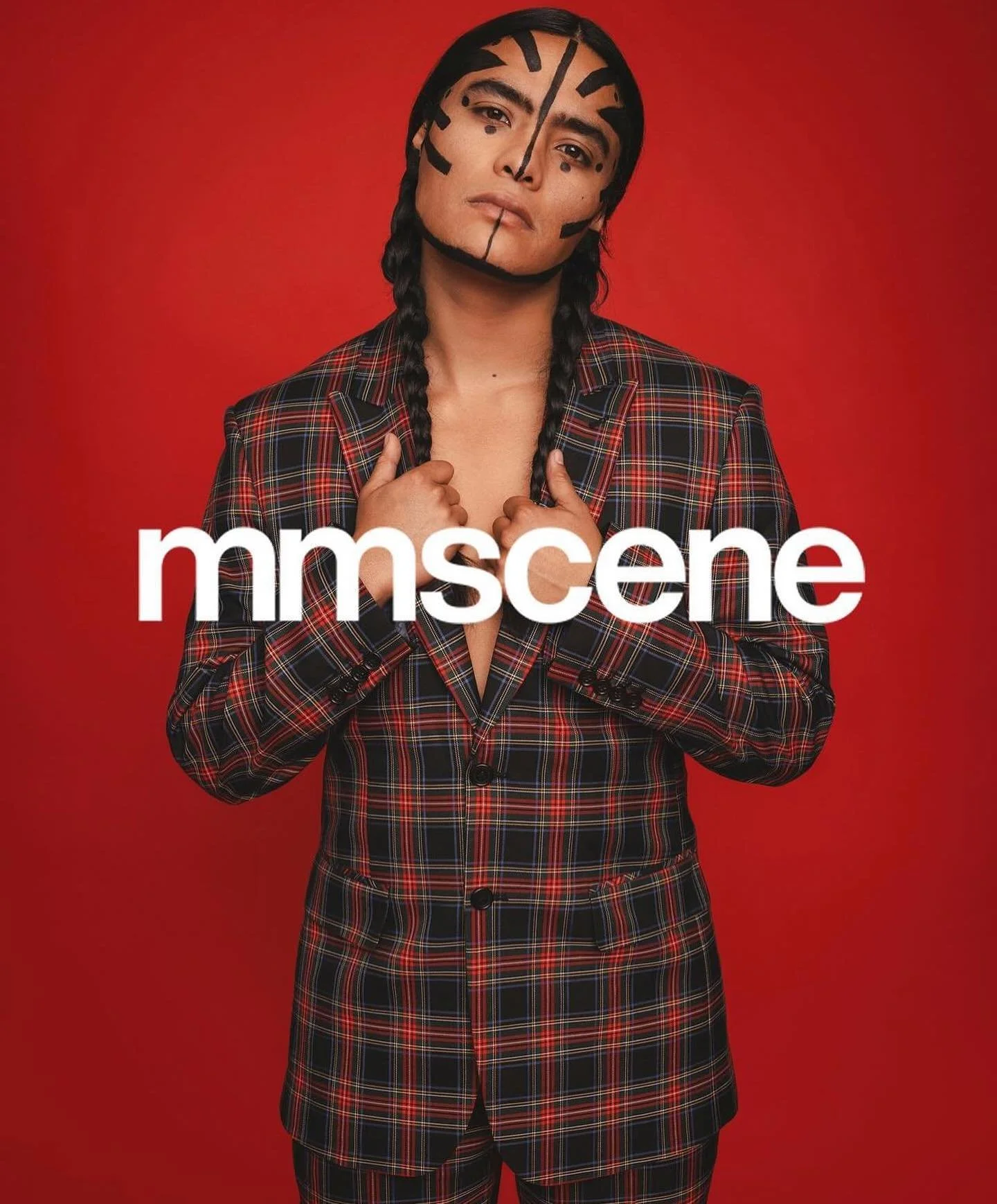 Person with long black hair and face paint resembling a zebra, wearing a plaid suit, standing against a red background, with the text 'mmscene' overlaid.