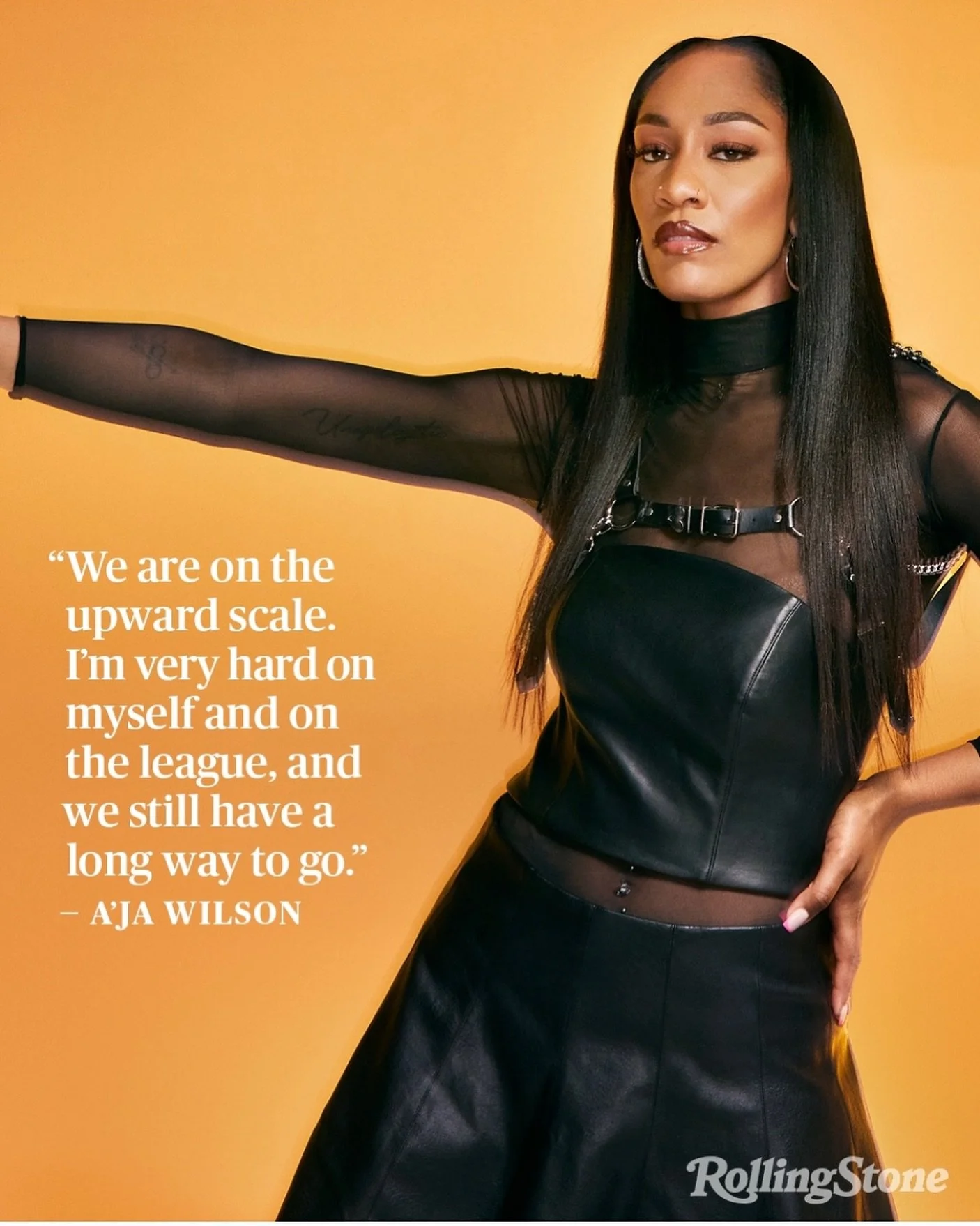 A woman with long, straight dark hair wearing a black leather top and sheer black long-sleeve shirt standing against a yellow background. She has her left hand on her hip and looks confidently at the camera. There is a quote on the left side of the i