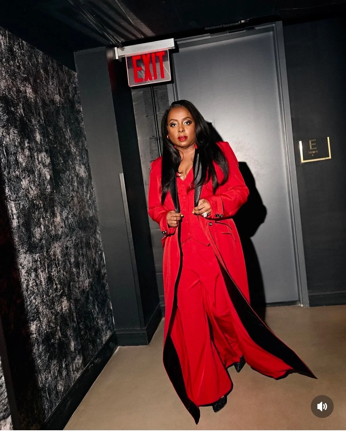 A woman with long black hair wearing a red coat and red pants, standing in front of a dark door with an exit sign above, in a modern interior space.