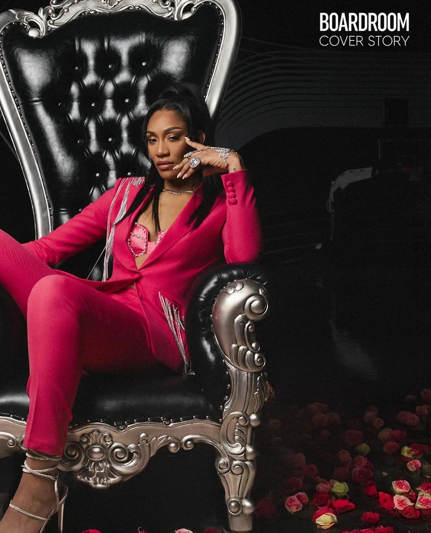 A woman in a pink suit sitting on an ornate black and silver throne surrounded by scattered rose petals.