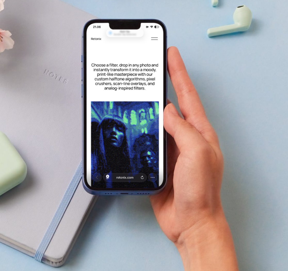 A person holding a smartphone with a website on the screen, showing a digital art filter applied to a photo of two women in a dark, neon-lit setting. The phone is held over a light blue surface with a closed gray notebook and a pair of light green wireless earbuds nearby.