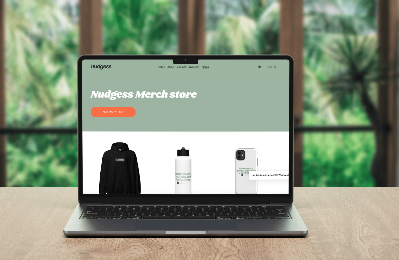 Laptop displaying the Nugdess merchandise store website with a green header, orange button, and images of a hoodie, water bottle, and smartphone on a wooden desk, with a blurred green outdoor background.