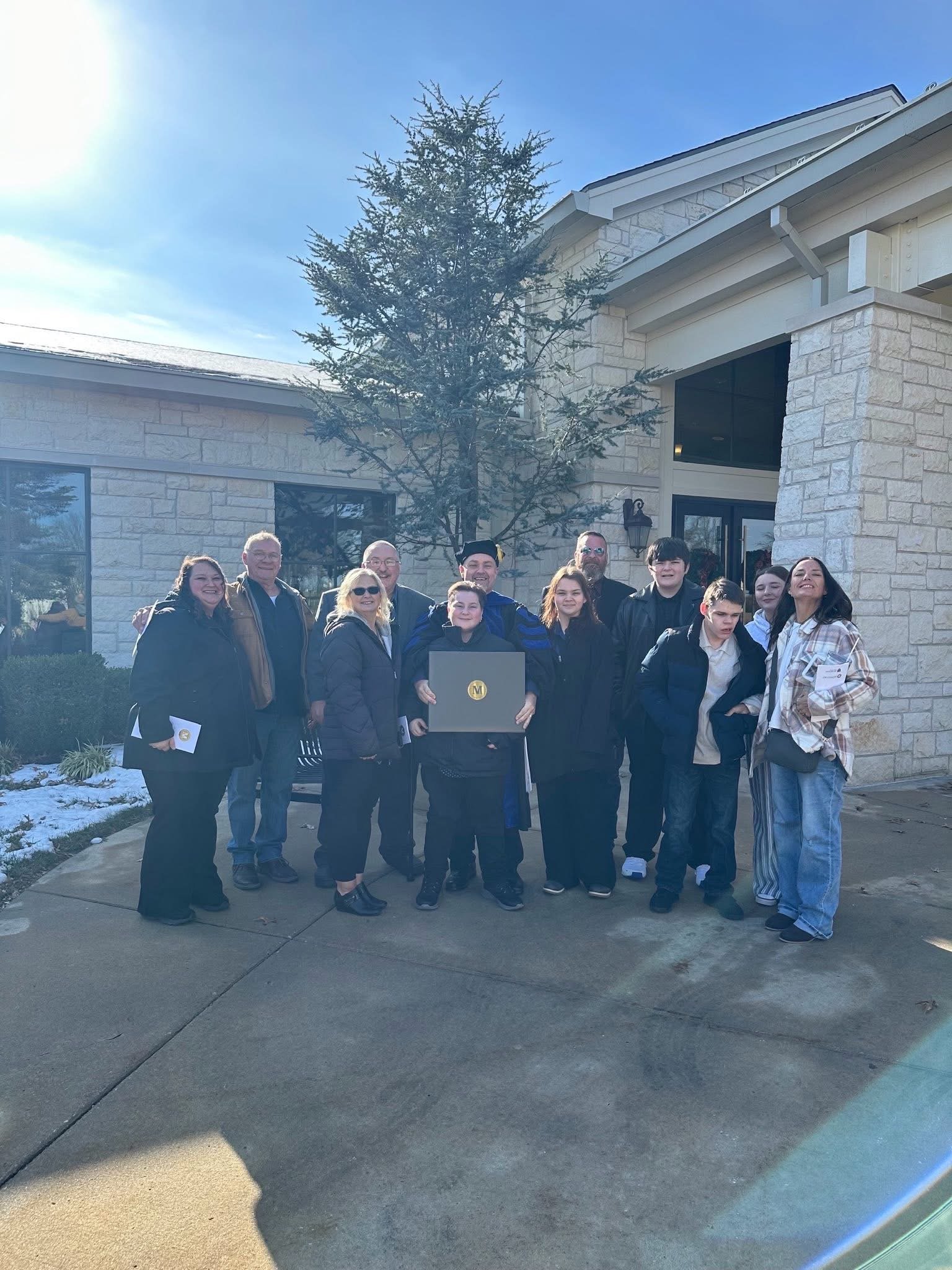 Group of twelve people standing outside a cream brick building on a concrete sidewalk, with snowy patches and a leafless tree in the background, during daylight.