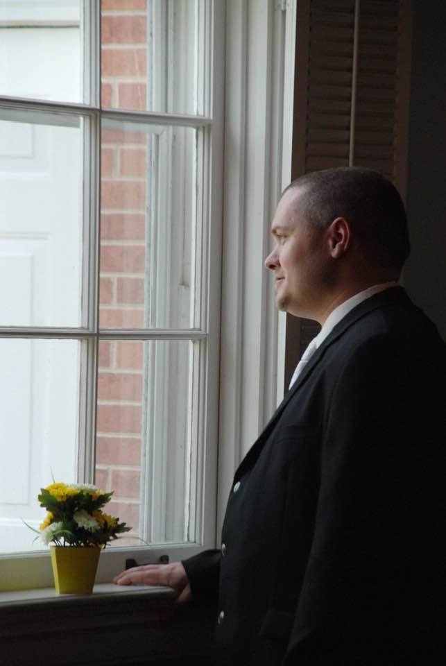 Man dressed in a formal black suit looking out of a window with a small yellow flower pot on the windowsill.