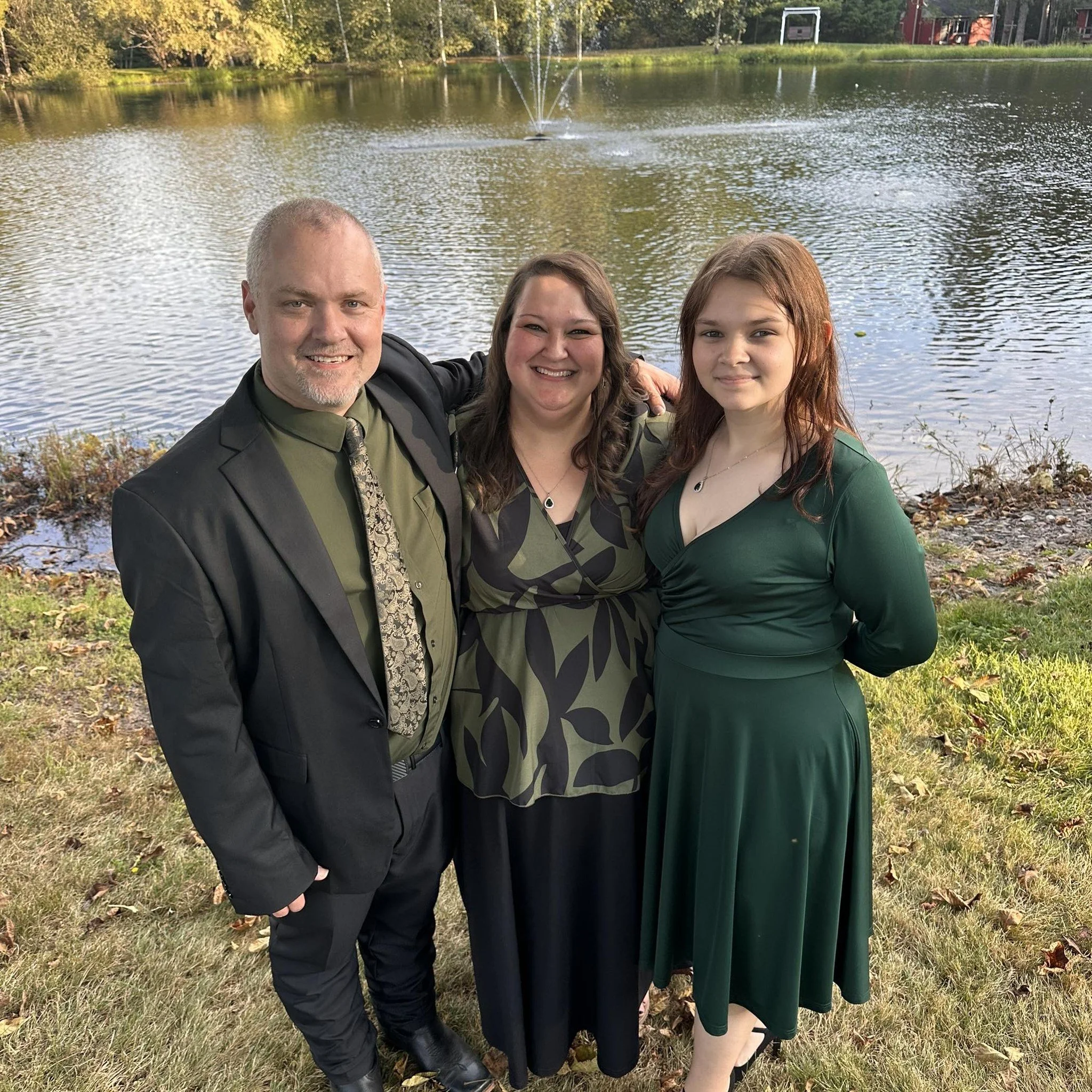 A family of three standing by a lake with trees and a fountain in the background. The man, woman, and teenage girl are smiling and dressed in green and black clothing.