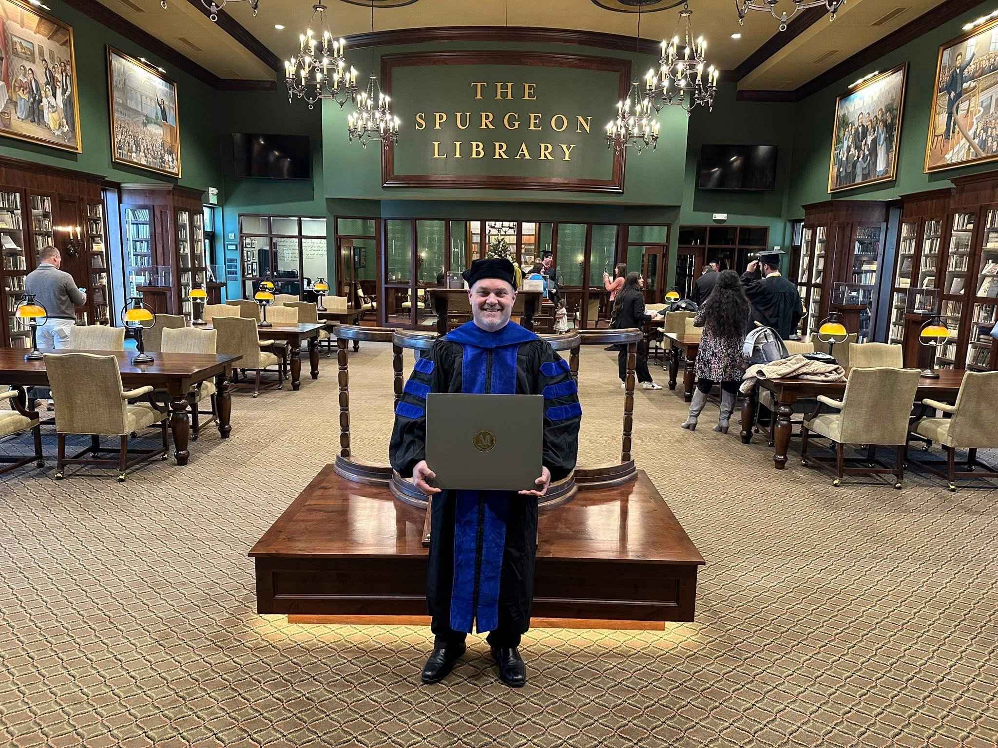 A person in academic regalia standing in the center of a library, holding a box with a gold emblem. The library has green walls, wood bookshelves, chandeliers, and framed paintings. There are other people in the background, some using laptops and others taking photos.
