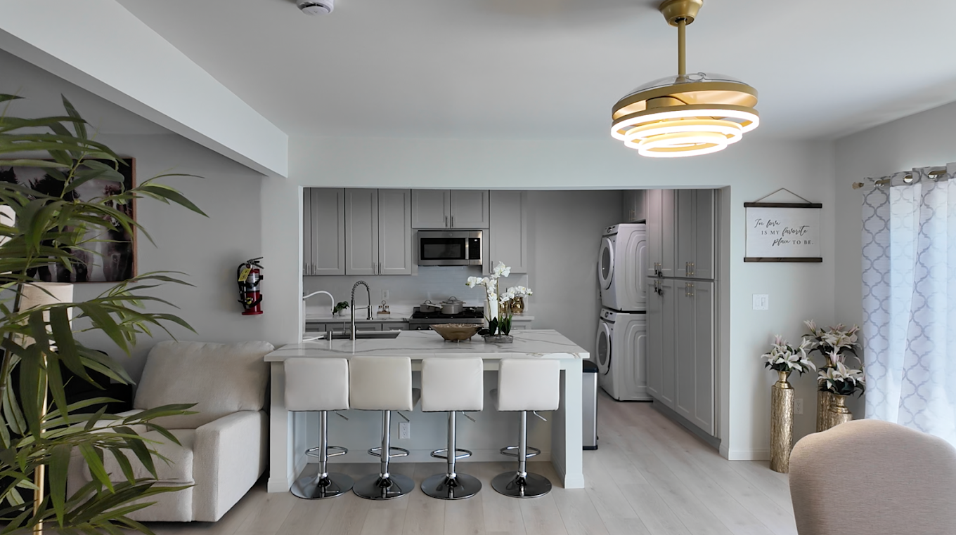 Modern kitchen with white cabinetry, marble island with four white chairs, stacked washer and dryer, and decorative elements like flowers and a wall sign.