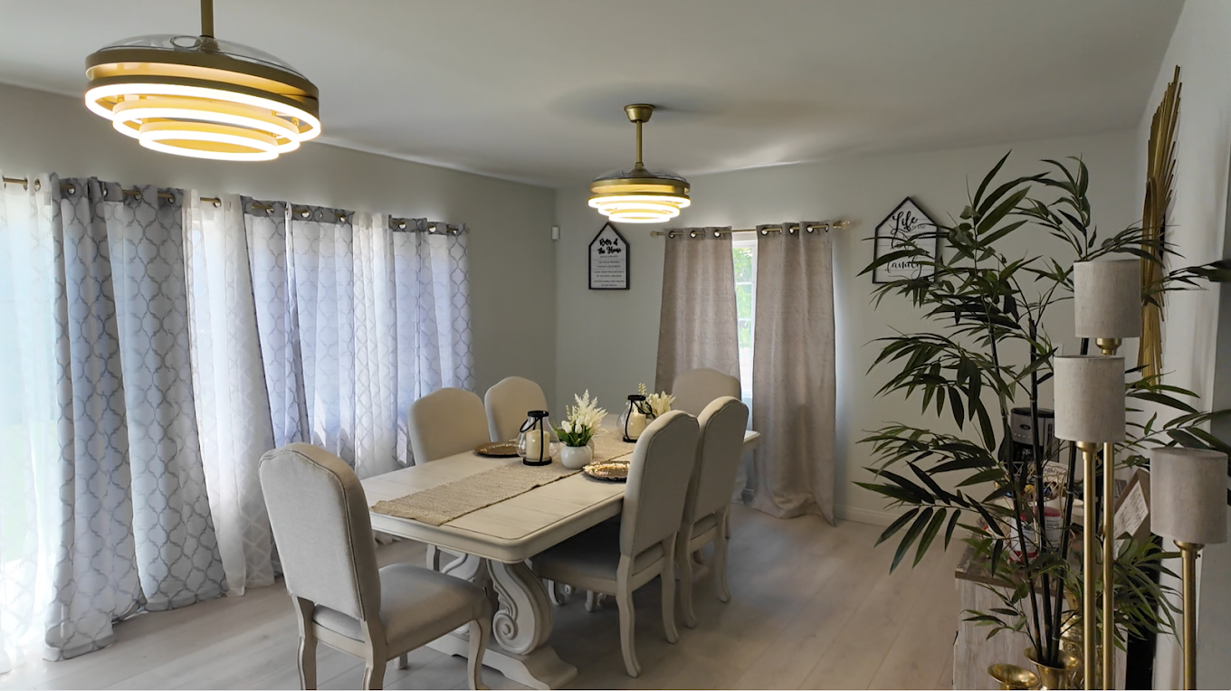 Dining room with a large table, six beige chairs, light-colored curtains, two modern chandelier lights, and a large potted plant.