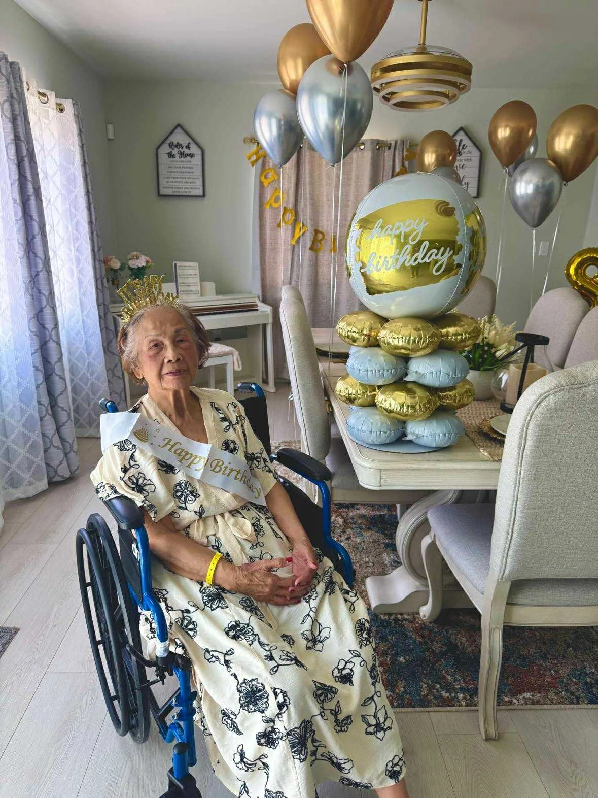 An elderly woman sitting in a wheelchair at a birthday celebration. She is wearing a dress with black floral patterns, a 'Happy Birthday' sash, and a gold crown. There are balloons and a decorative cake in the background, with a 'Happy Birthday' banner hanging across the room.