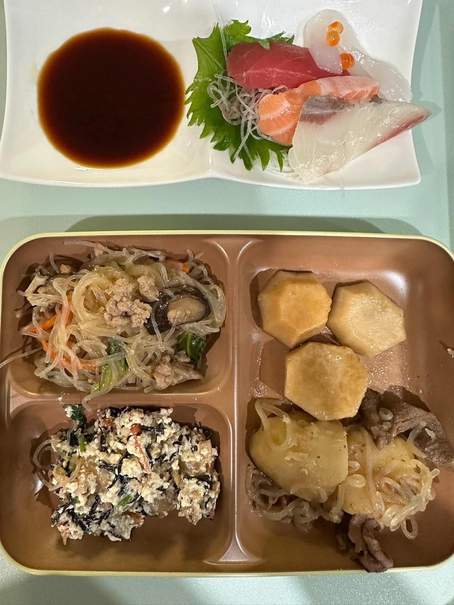A tray with four compartments containing Japanese food, including cooked vegetables and tofu, with sashimi and soy sauce on a plate.