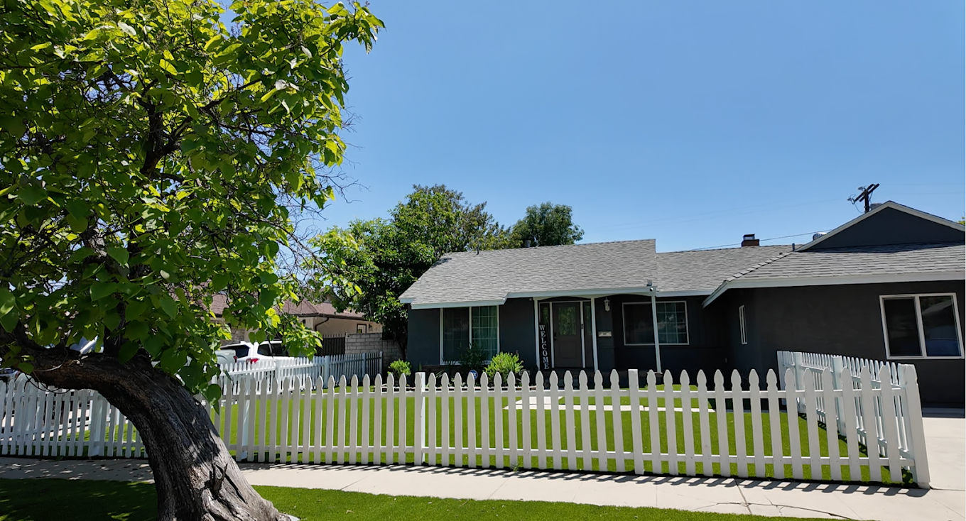 View of a gray house with a white picket fence and a lush green lawn, with a large tree on the left and a clear blue sky.