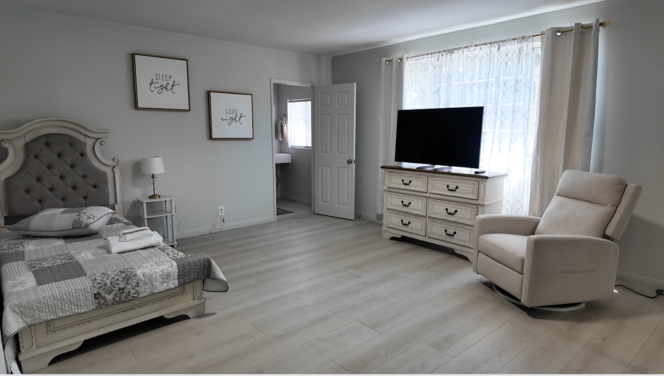 A bedroom with a bed, a nightstand, a lamp, wall art reading 'Sleep tight' and 'Good night', a television on a dresser, an armchair near the window, and a view of a small bathroom with a sink.