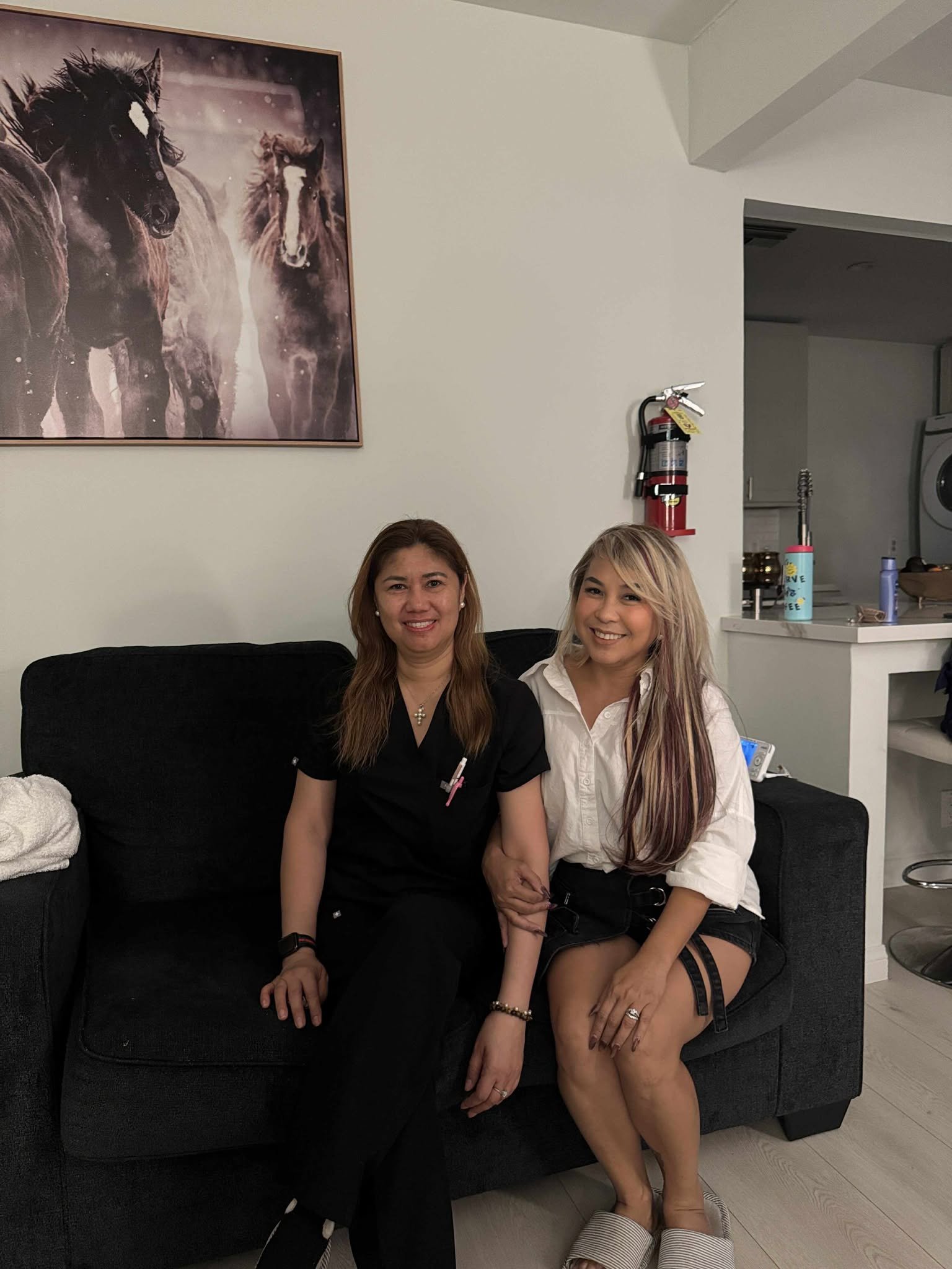 Two women sitting together on a black couch in a living room, smiling at the camera. One is wearing a black top, and the other is wearing a white shirt with black shorts. There is a framed picture of horses on the wall behind them, and a fire extinguisher is mounted on the wall nearby.