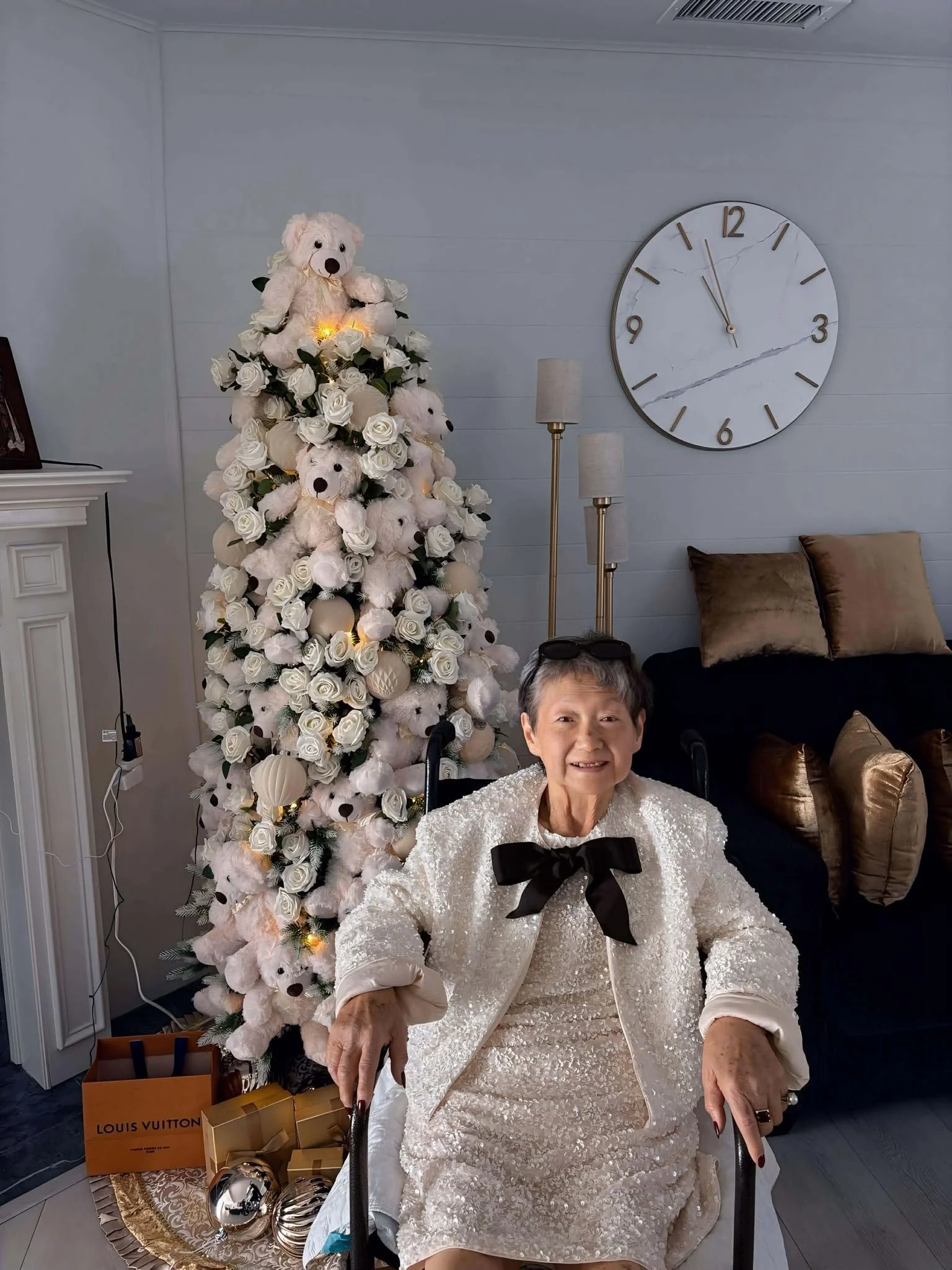 An elderly woman sitting in a chair in a living room decorated for Christmas with a white Christmas tree, gold and cream pillows, a clock showing 12:07, and festive ornaments.