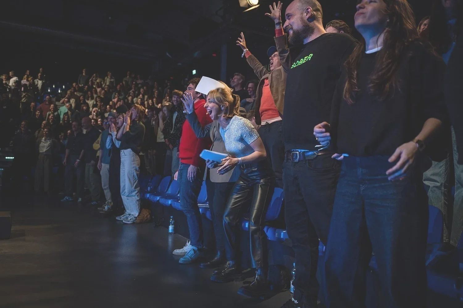 Menschen jubeln und feiern bei einer Veranstaltung in einem dunklen Raum, während sie auf einer Bühne stehen.