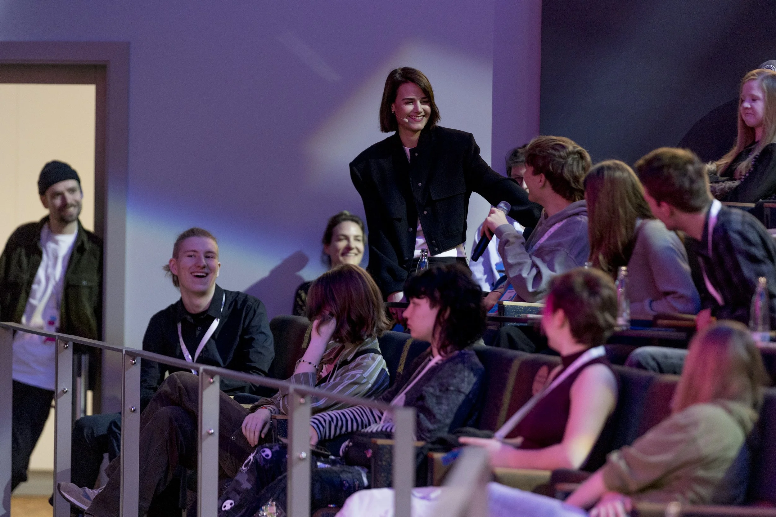 Junge Frau mit Mikrofon, die in einem Theater- oder Konferenzraum mit Publikum spricht, während Teilnehmer im Publikum lachen und sich unterhalten.