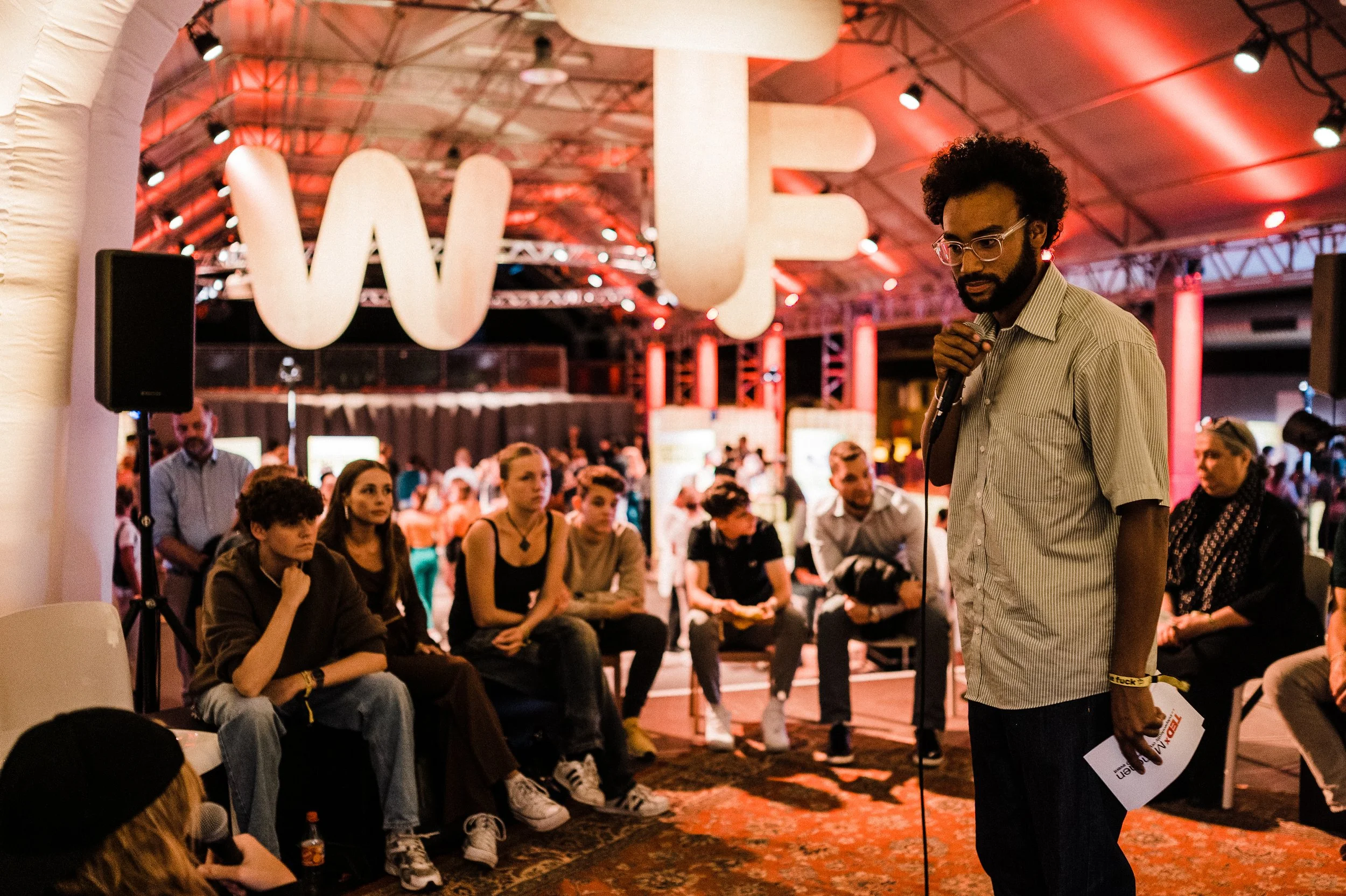 Ein Mann hält ein Mikrofon und spricht zu einem Publikum bei einer Veranstaltung. Es sitzen mehrere Menschen und hören zu, im Hintergrund sind große weiße Buchstaben und eine große Struktur.