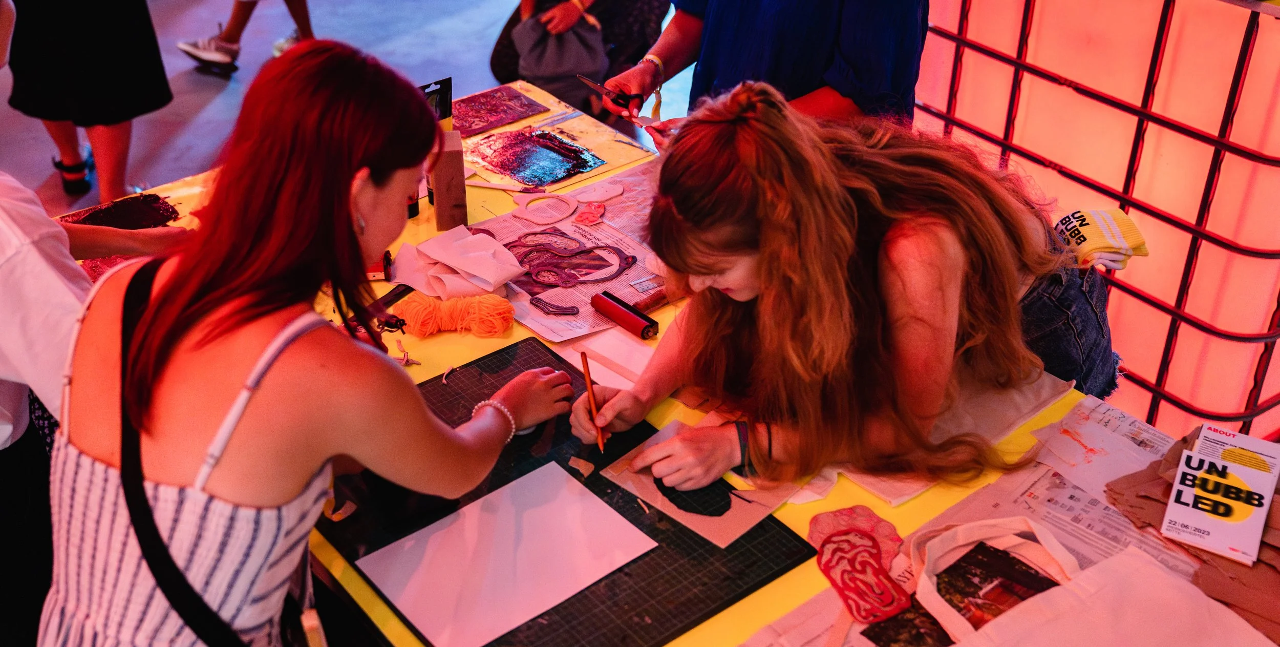 Zwei Frauen arbeiten an einem Basteltisch mit verschiedenen Materialien und Werkzeugen, umgeben von bunten Papieren und Verpackungen.