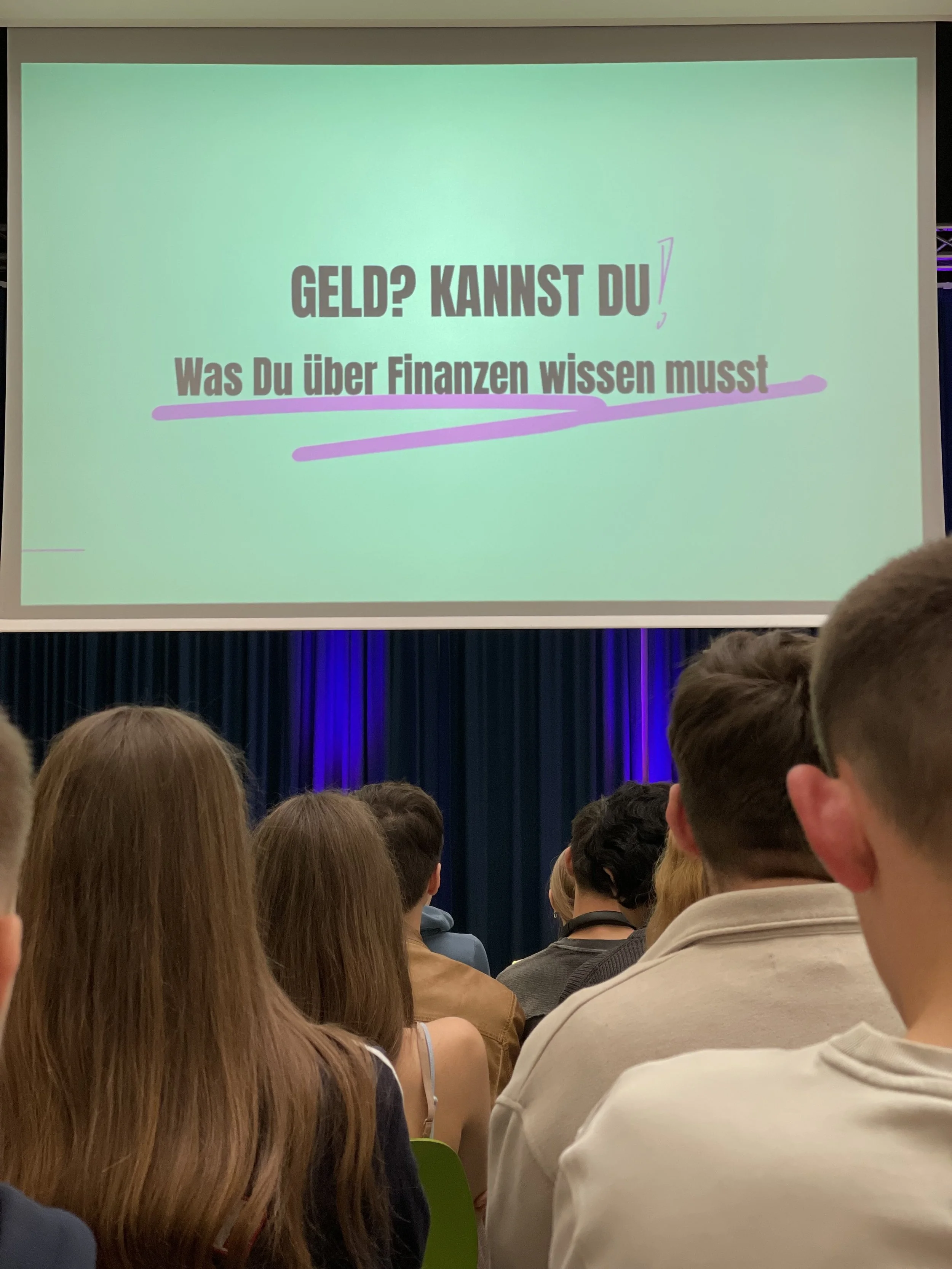 Ein Publikum sitzt vor einer großen Leinwand, auf der ein Präsentationsbild mit dem deutschen Text 'GELD? KANNST DU! Was Du über Finanzen wissen musst' projiziert wird.