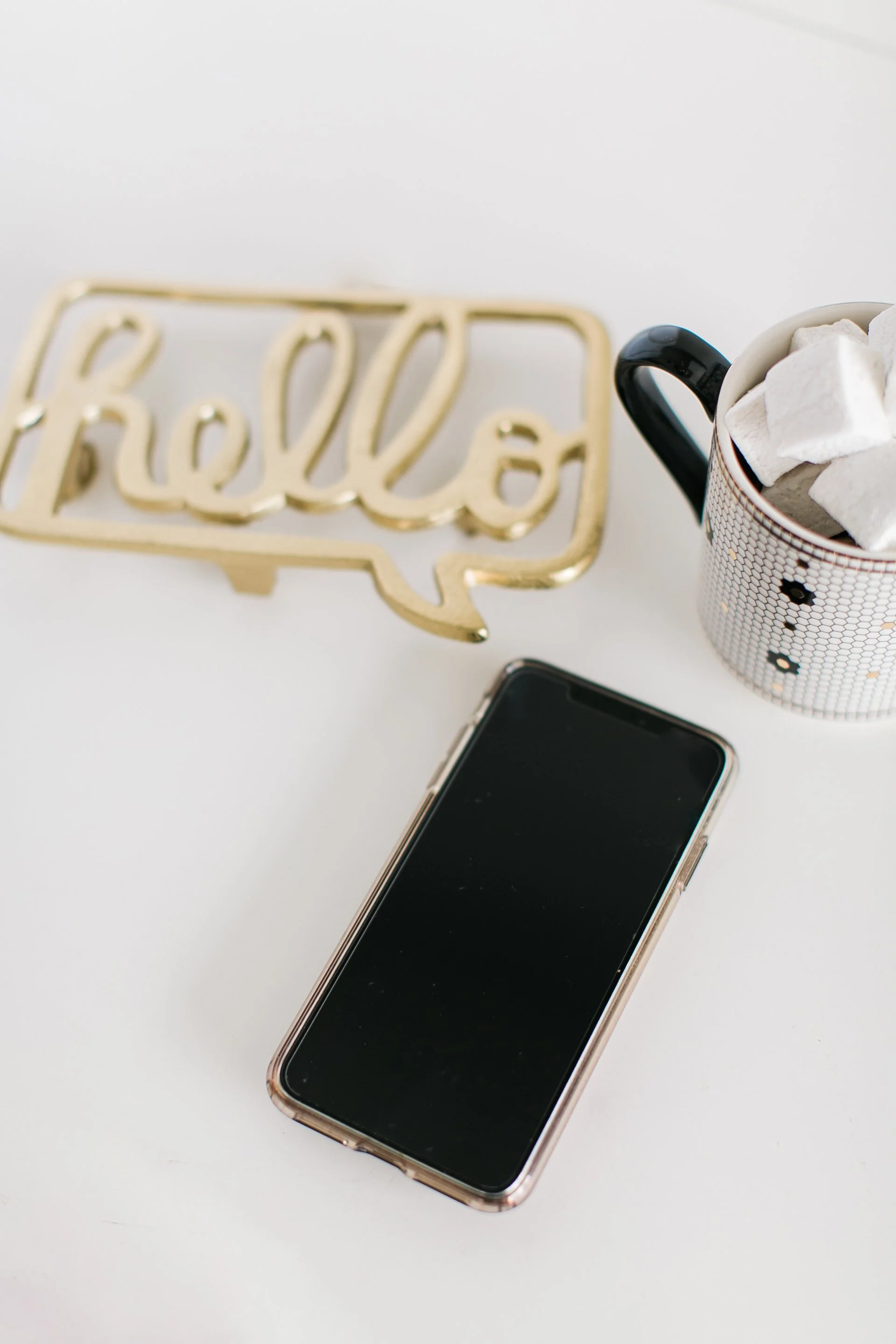 A smartphone, a mug with marshmallows, and a gold decorative sign that says 'Hello' on a white surface.