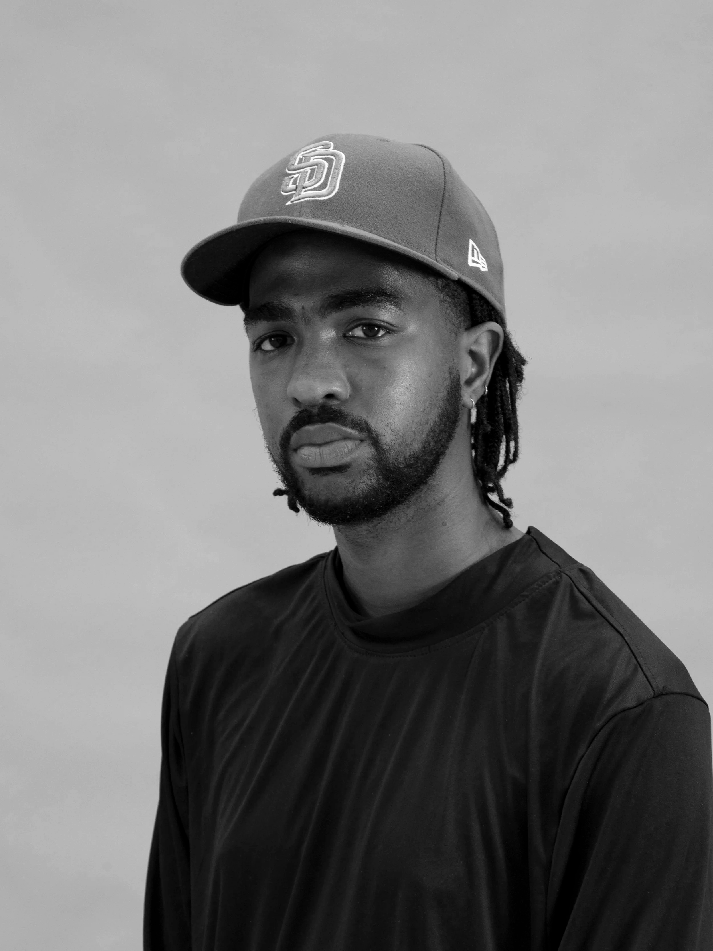 Black and white photo of a man with dreadlocks wearing a San Diego Padres baseball cap and a black shirt, standing against a plain wall.