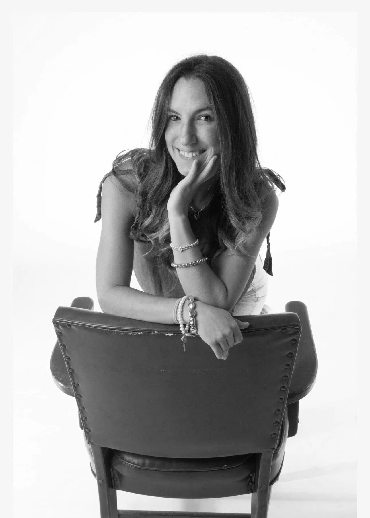 Black and white portrait of a young woman sitting on a chair with a smile, resting her chin on her hand, wearing bracelets and a sleeveless top.
