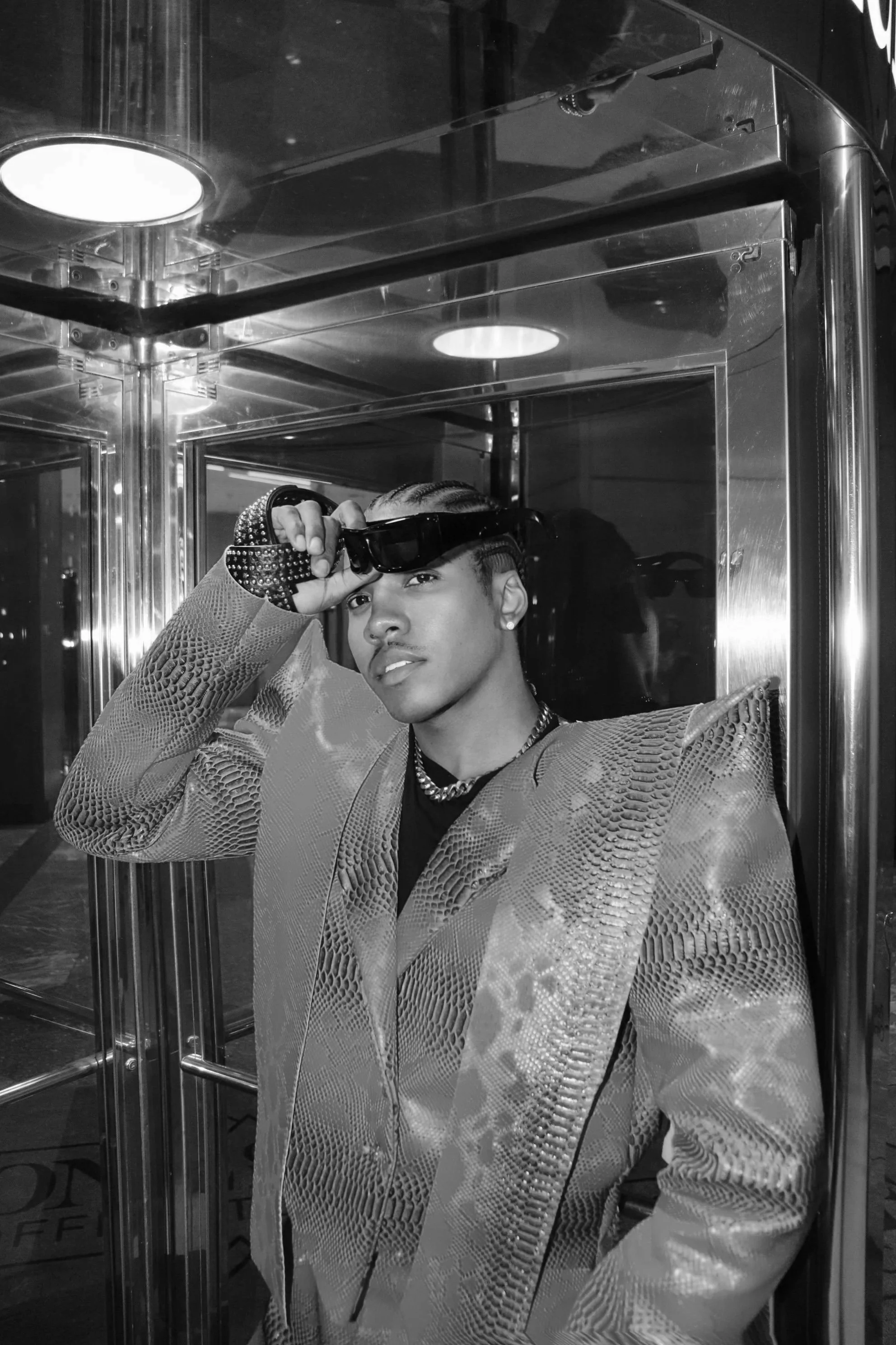A young person with styled hair and earrings taking a selfie in a mirror inside an elevator with reflective walls. They are wearing a textured, patterned jacket and holding a pair of sunglasses to their forehead.