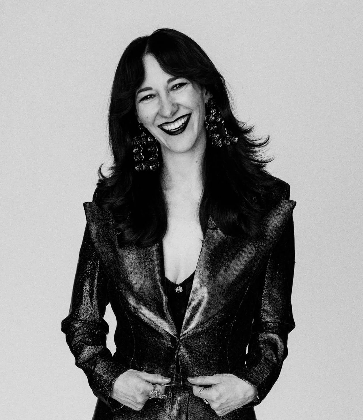 Black and white portrait of a woman smiling, wearing a shiny leather jacket, large earrings, and styled hair, standing against a plain background.