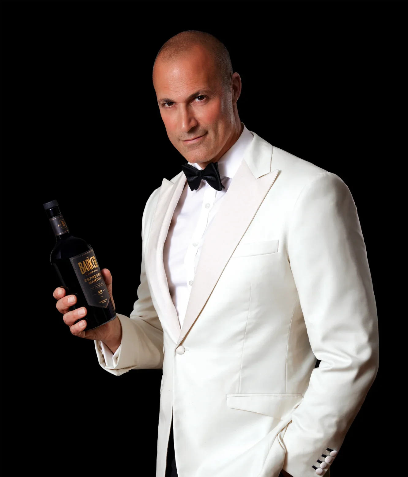 A man wearing a white tuxedo with a black bow tie holding a bottle of BARKER Espresso Martini against a black background.