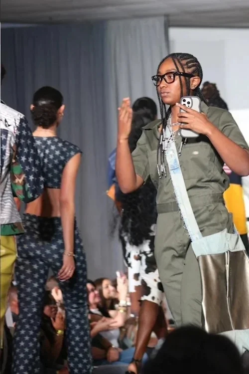 Young girl with braids, glasses, and military-style clothing taking a photo with her phone at a fashion show.