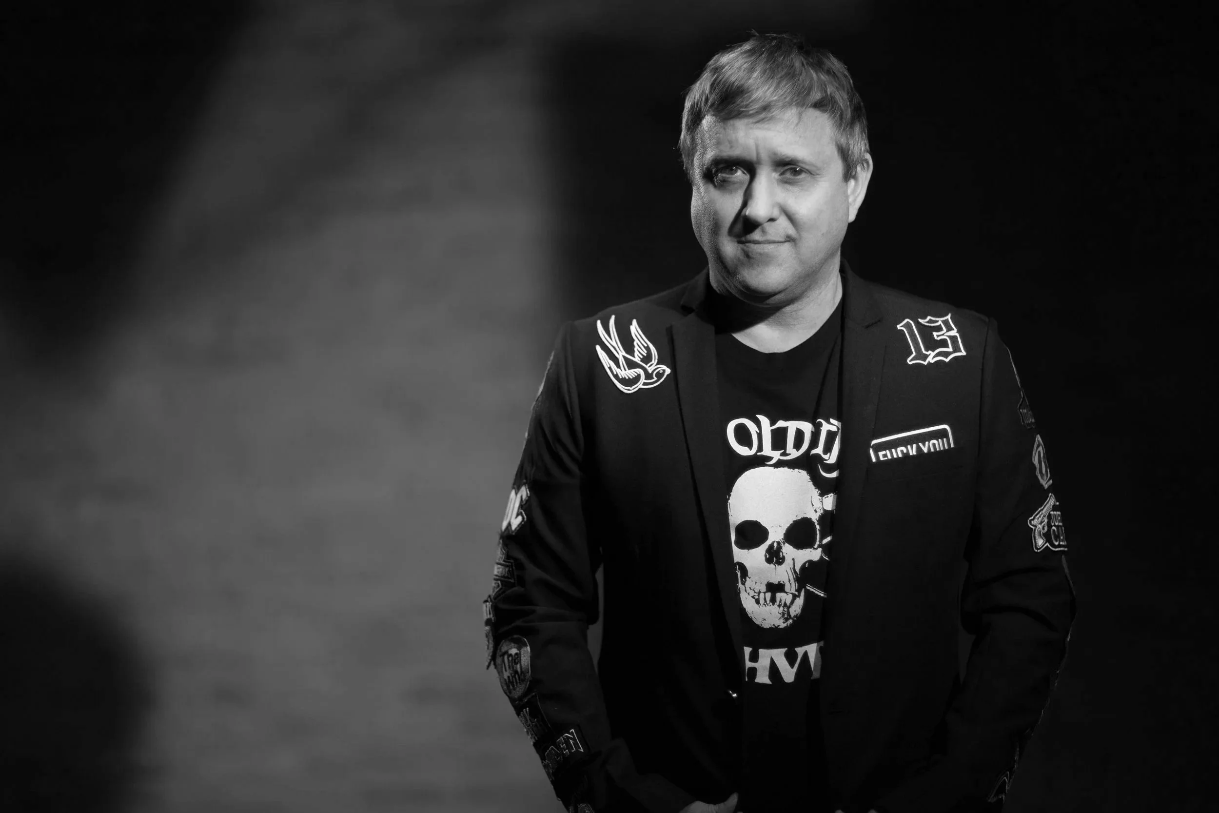 Black and white photograph of a man with short hair, wearing a jacket with patches and a skull graphic t-shirt, standing against a dark background. The man is smiling slightly and looking towards the camera.