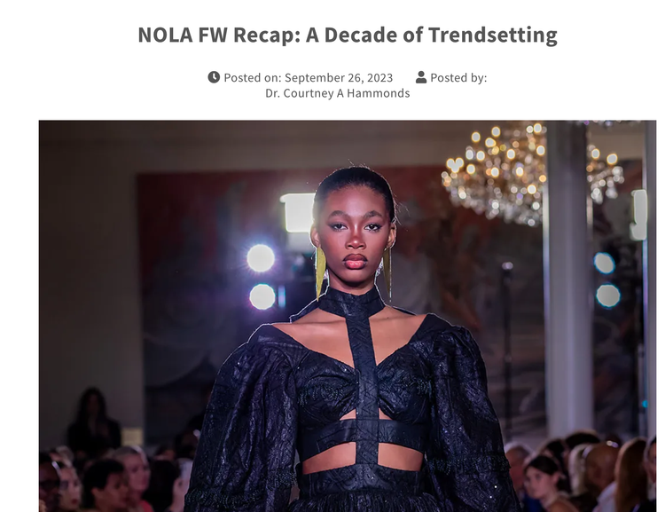 A female model walking the runway at a fashion show, wearing a dark, Gothic-inspired outfit with cutouts and puffed sleeves, with bright lights and chandeliers in the background, and an audience watching.