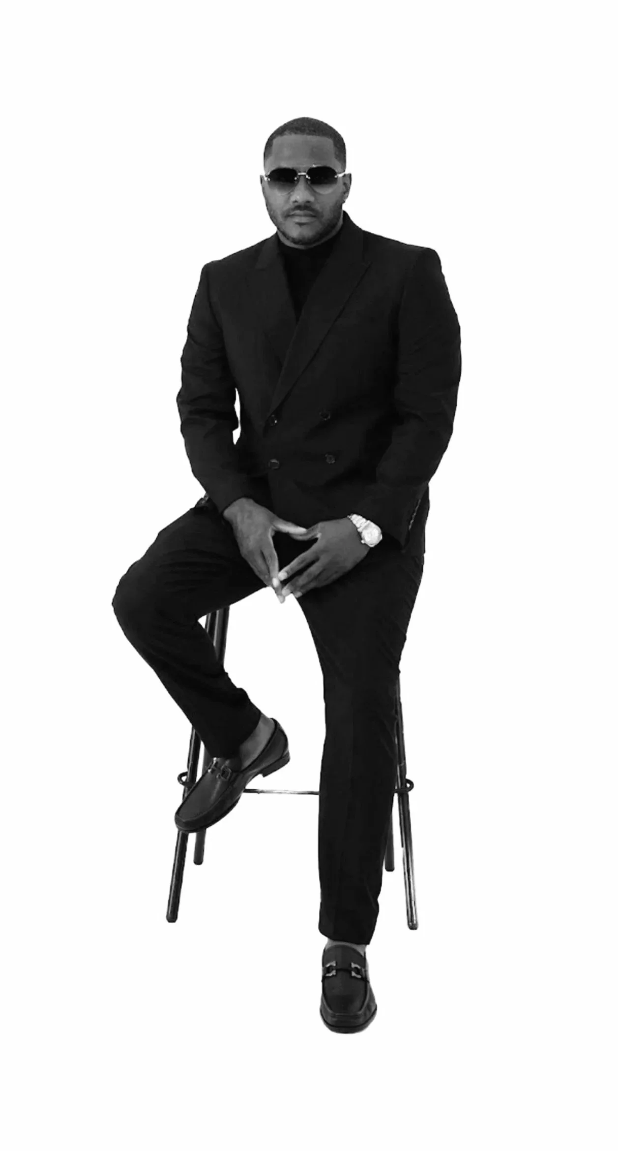 A man in a black suit with sunglasses, sitting on a stool with one leg crossed over the other, against a plain white background.
