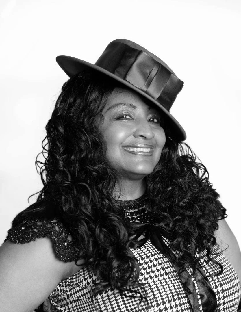 Black and white photo of a woman smiling, wearing a patterned top, with curly hair, and a wide-brimmed hat tilted to the side.