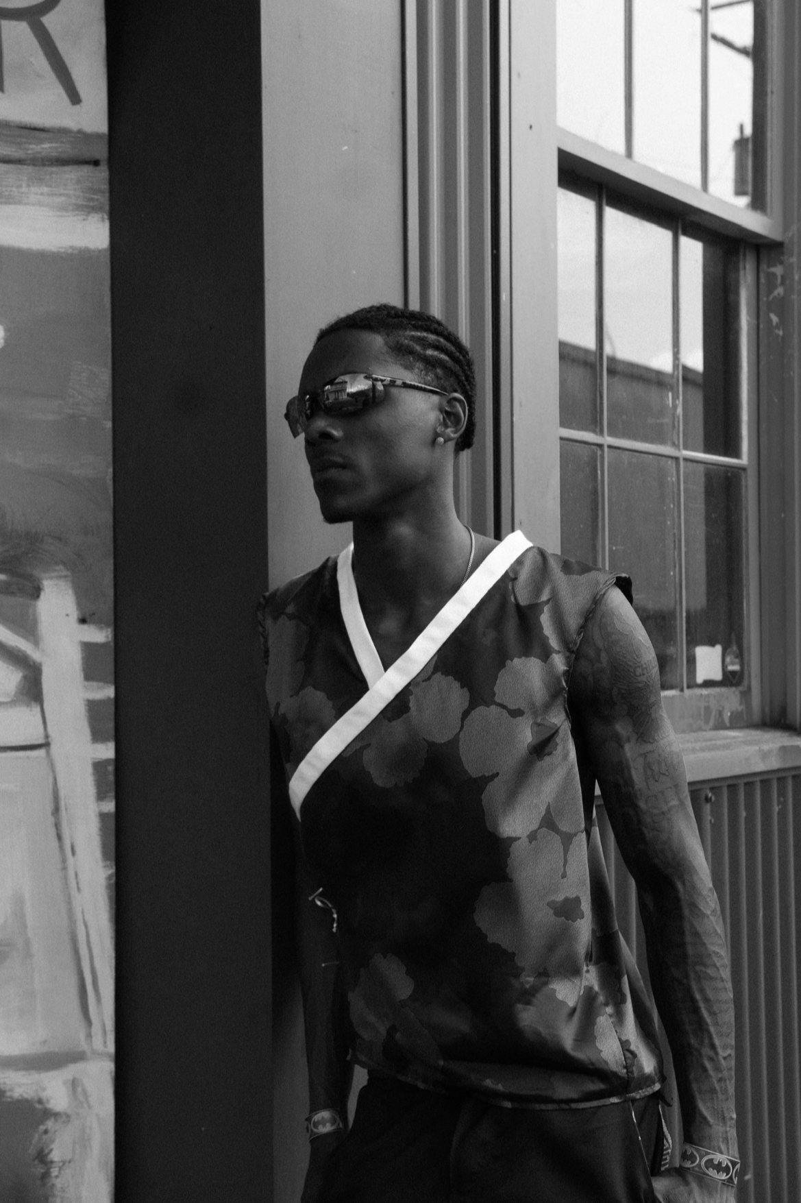 A man with braided hair wearing sunglasses and a sleeveless camouflage top standing near a window in an urban setting.