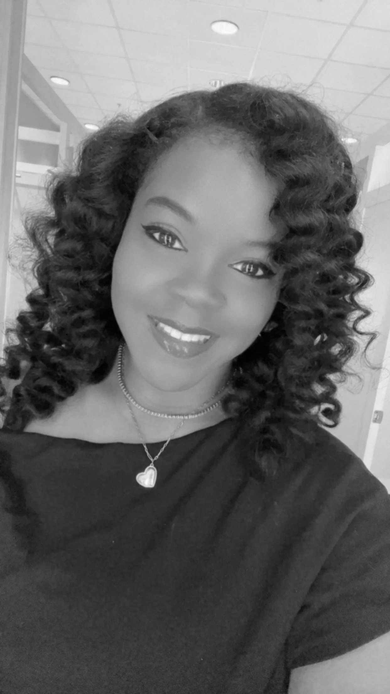 A woman with curly hair, smiling, wearing a heart-shaped necklace and a dark top, indoors.